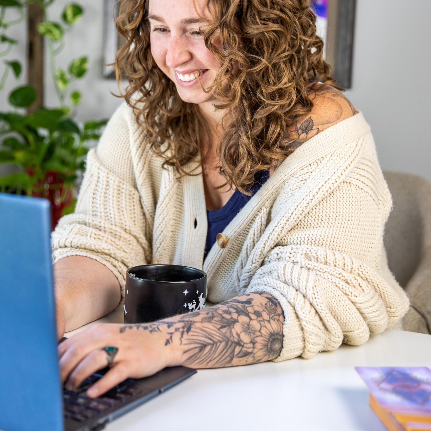 A woman with curly hair and a cream cardigan typing on her laptop, with a mug of coffee nearby.