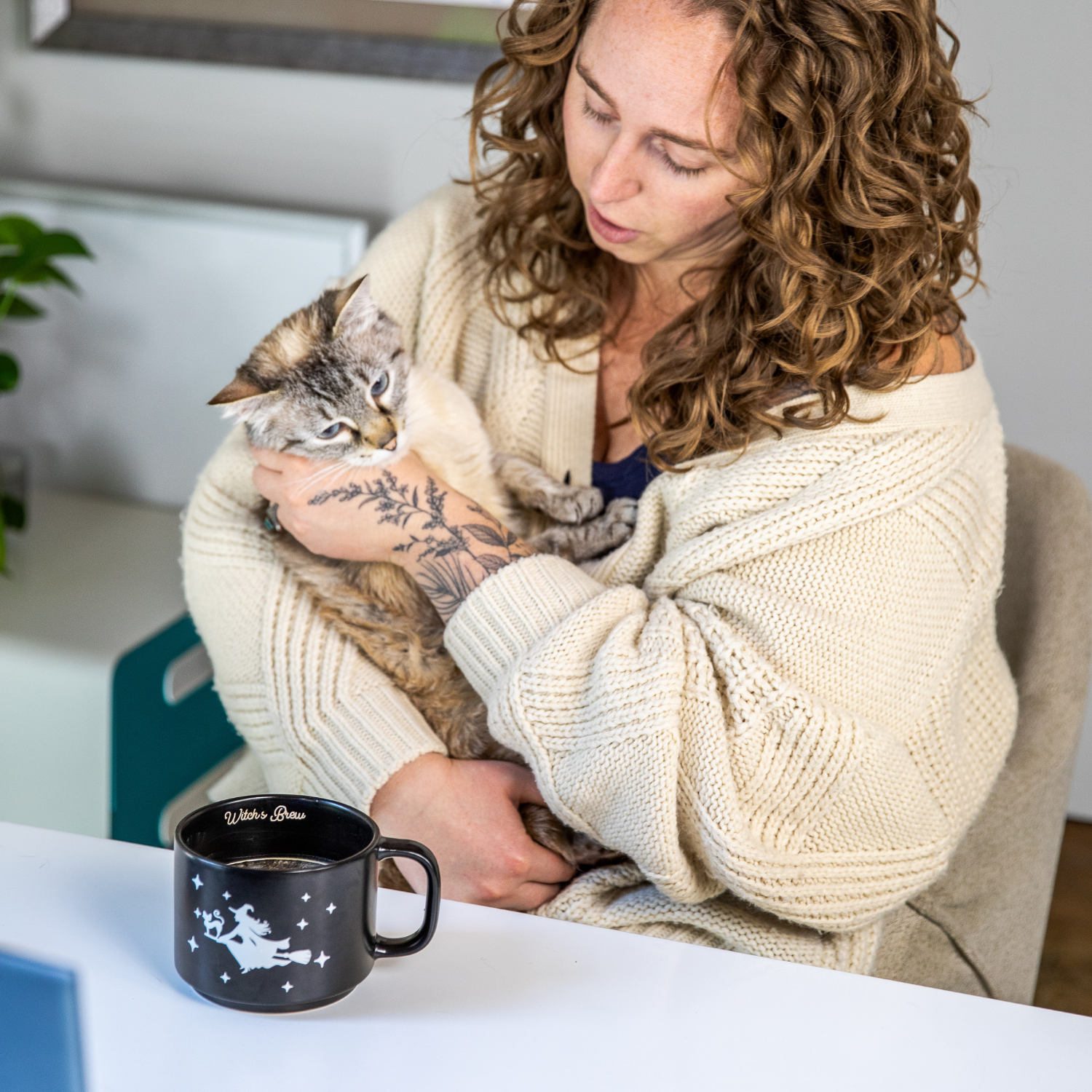 A woman with curly hair and a cream cardigan cuddling a tabby cat, with a mug of coffee nearby.