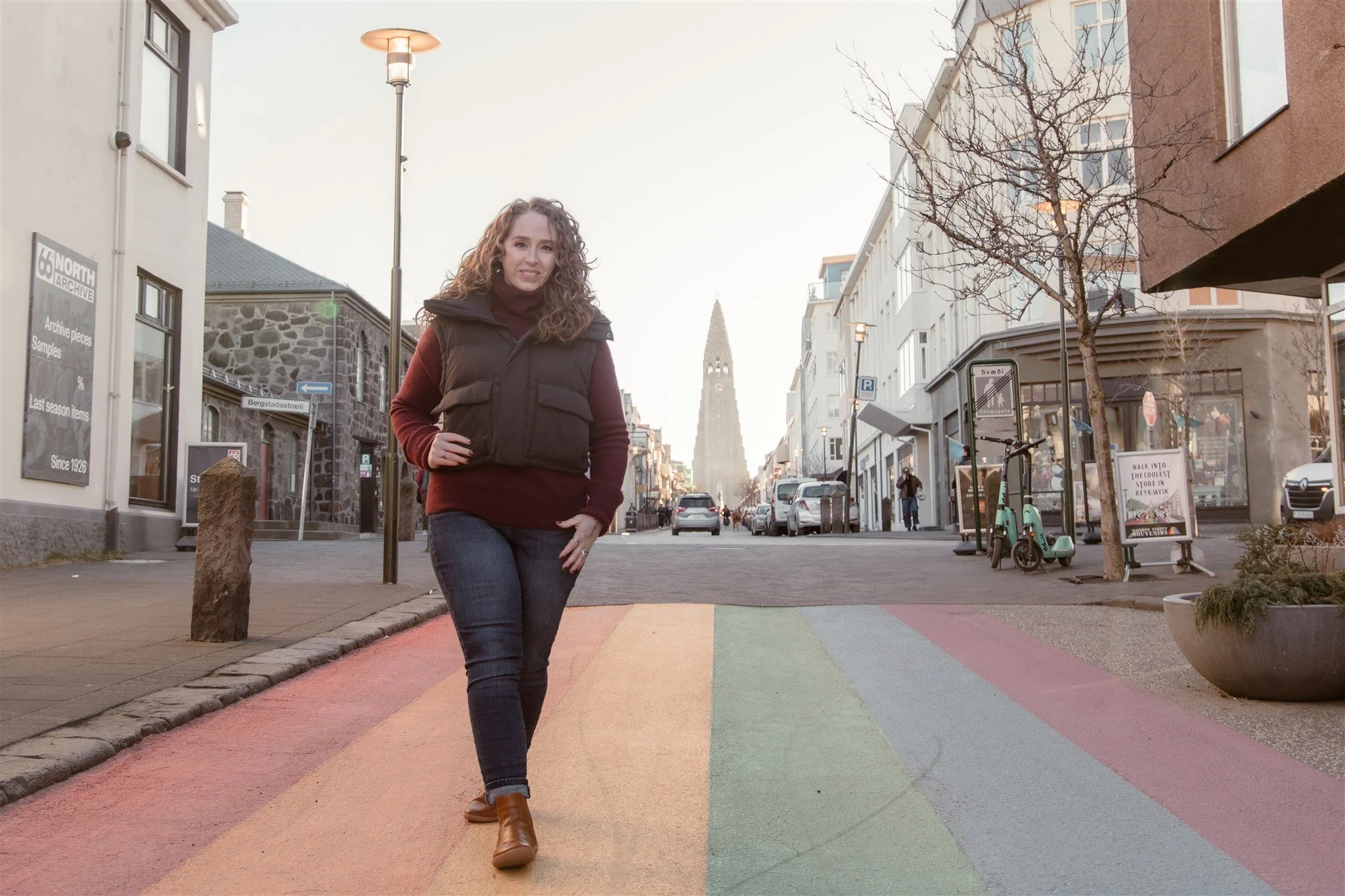 Woman walking on a rainbow-colored crosswalk in an urban setting with buildings, trees, parked bicycles, and a clock tower in the background.
