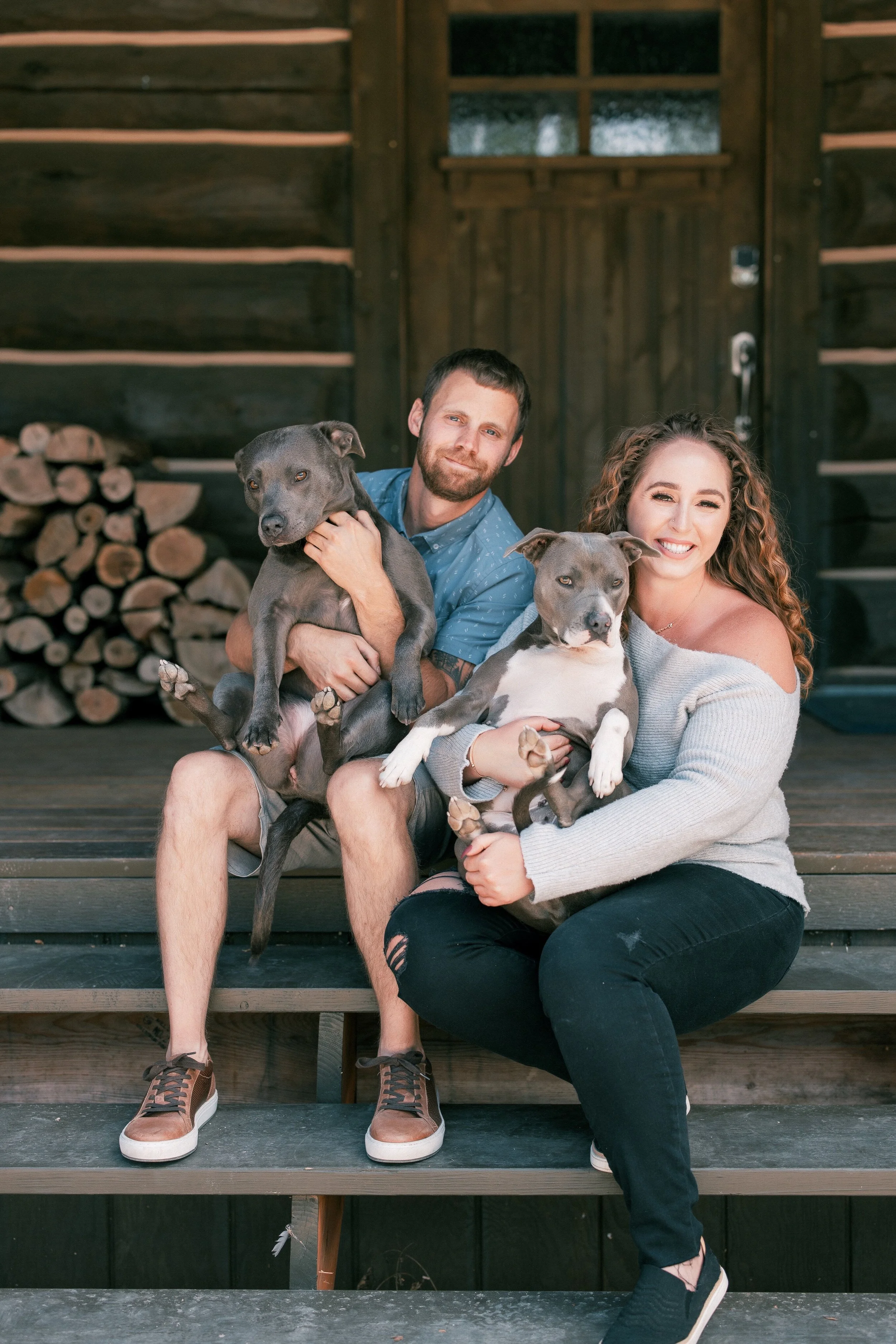A young man with short dark hair and a beard, and a young woman with curly brown hair, sitting on wooden steps outside a rustic dark wood building, each holding a gray and white dog.