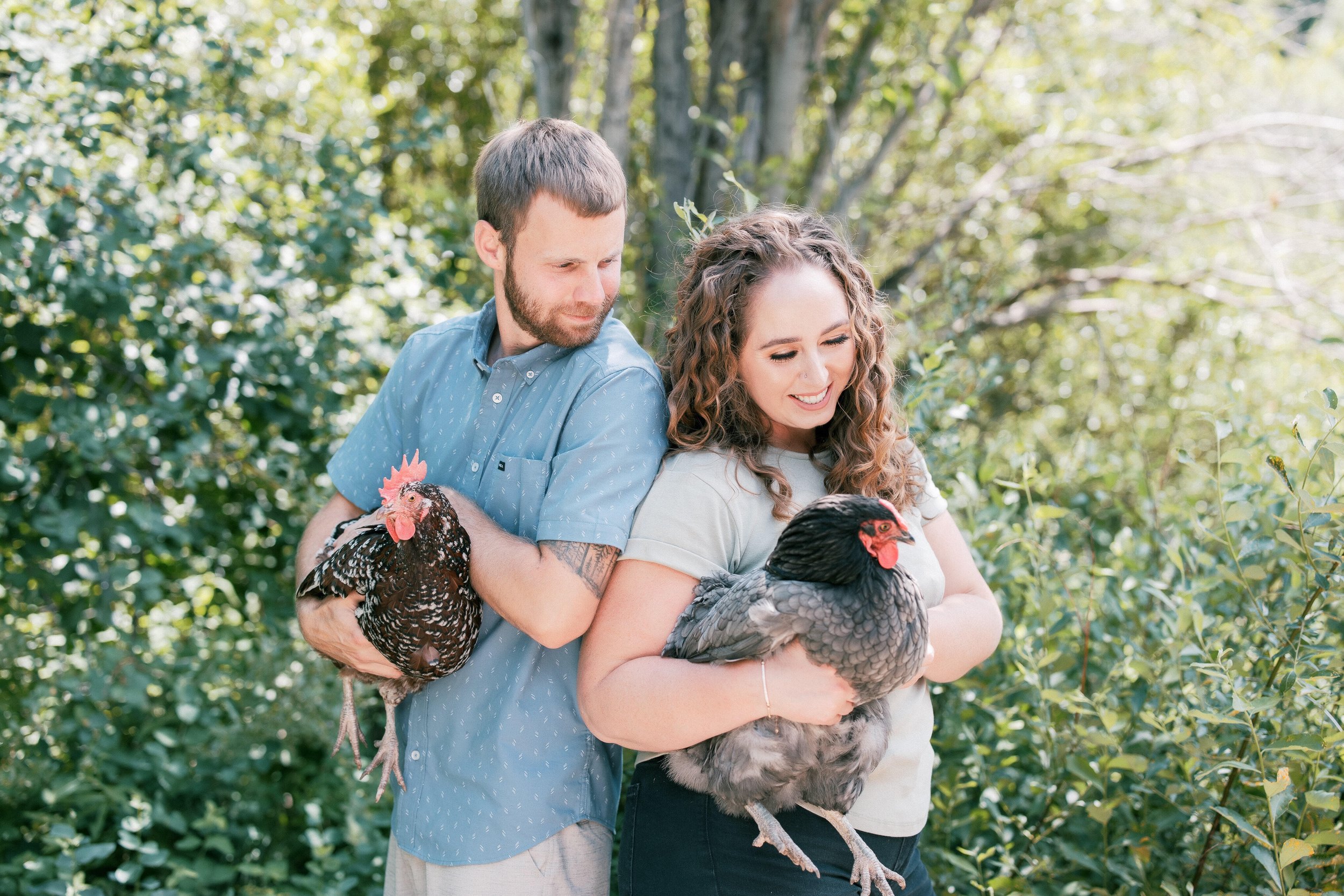 A man and a woman holding large turkeys outdoors in a lush green wooded area, smiling and looking at the turkeys.