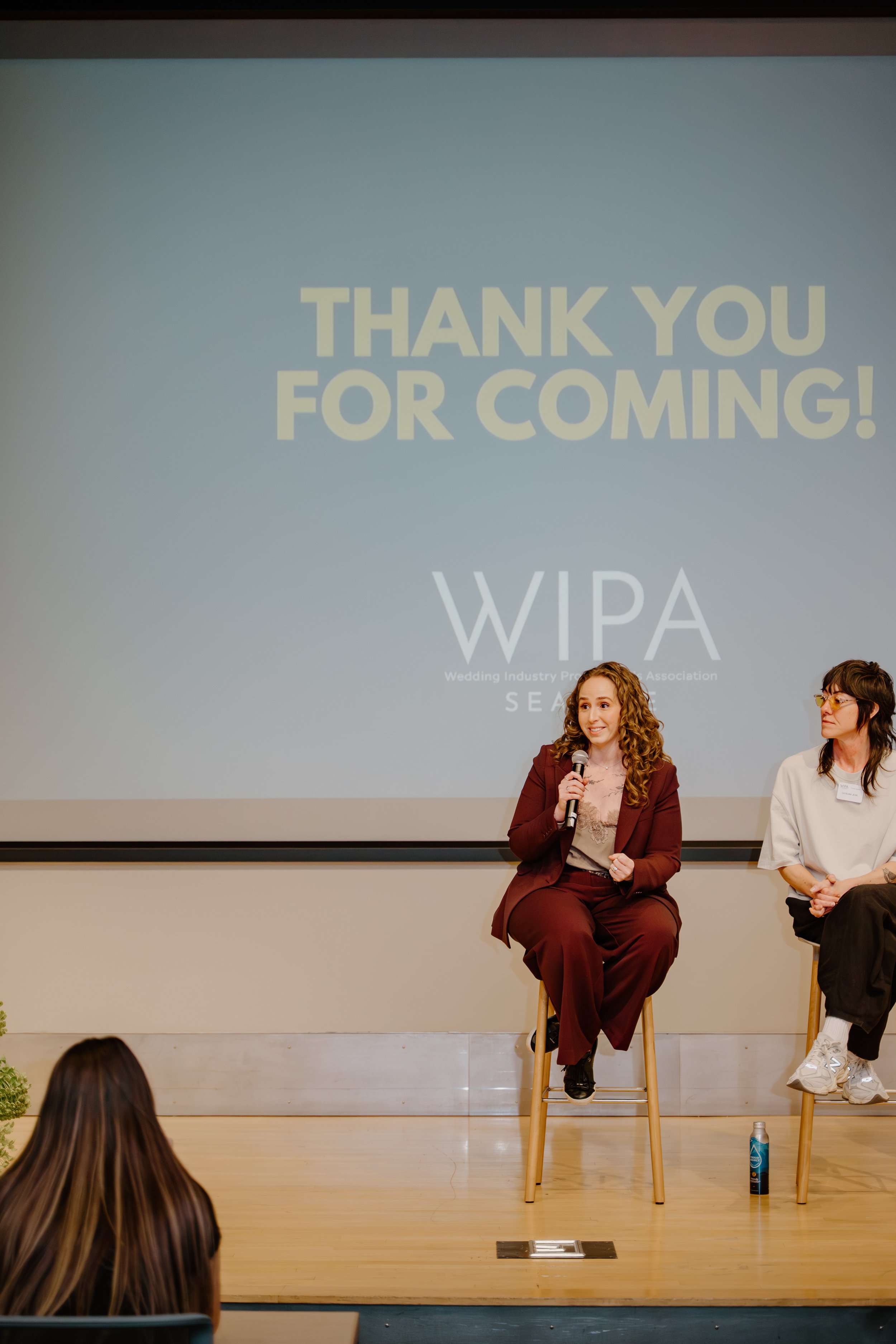 A woman with curly hair and a suit sitting on a barstool, holding a microphone, speaking at an event. Behind her is a screen displaying the words 'Thank You For Coming!' and 'WIPA' with additional smaller text. Another woman is sitting next to her. Audience members are visible in the foreground.