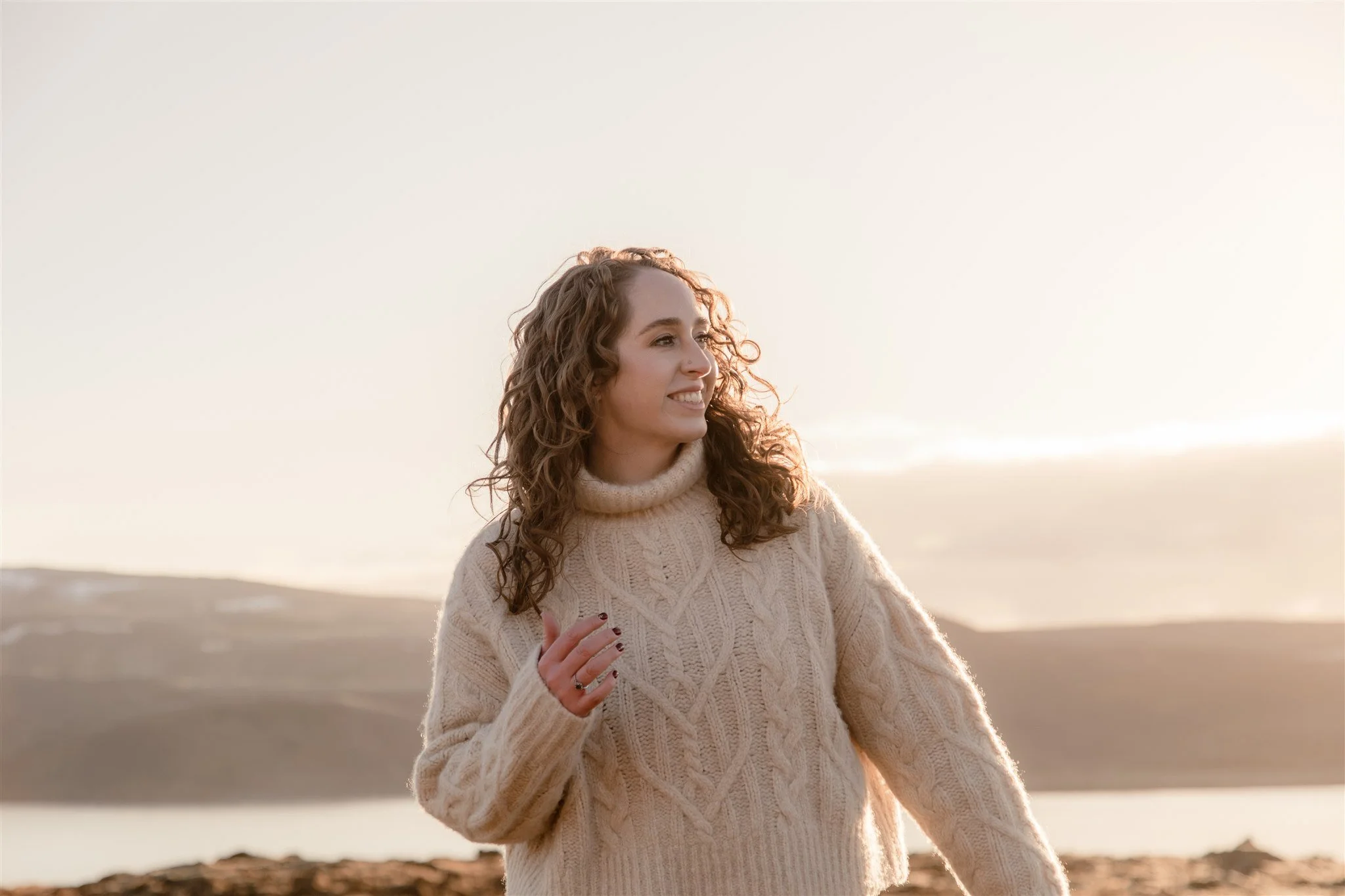 A woman with curly hair smiling and looking to the side, wearing a beige knit sweater, outdoors on a beach at sunset.