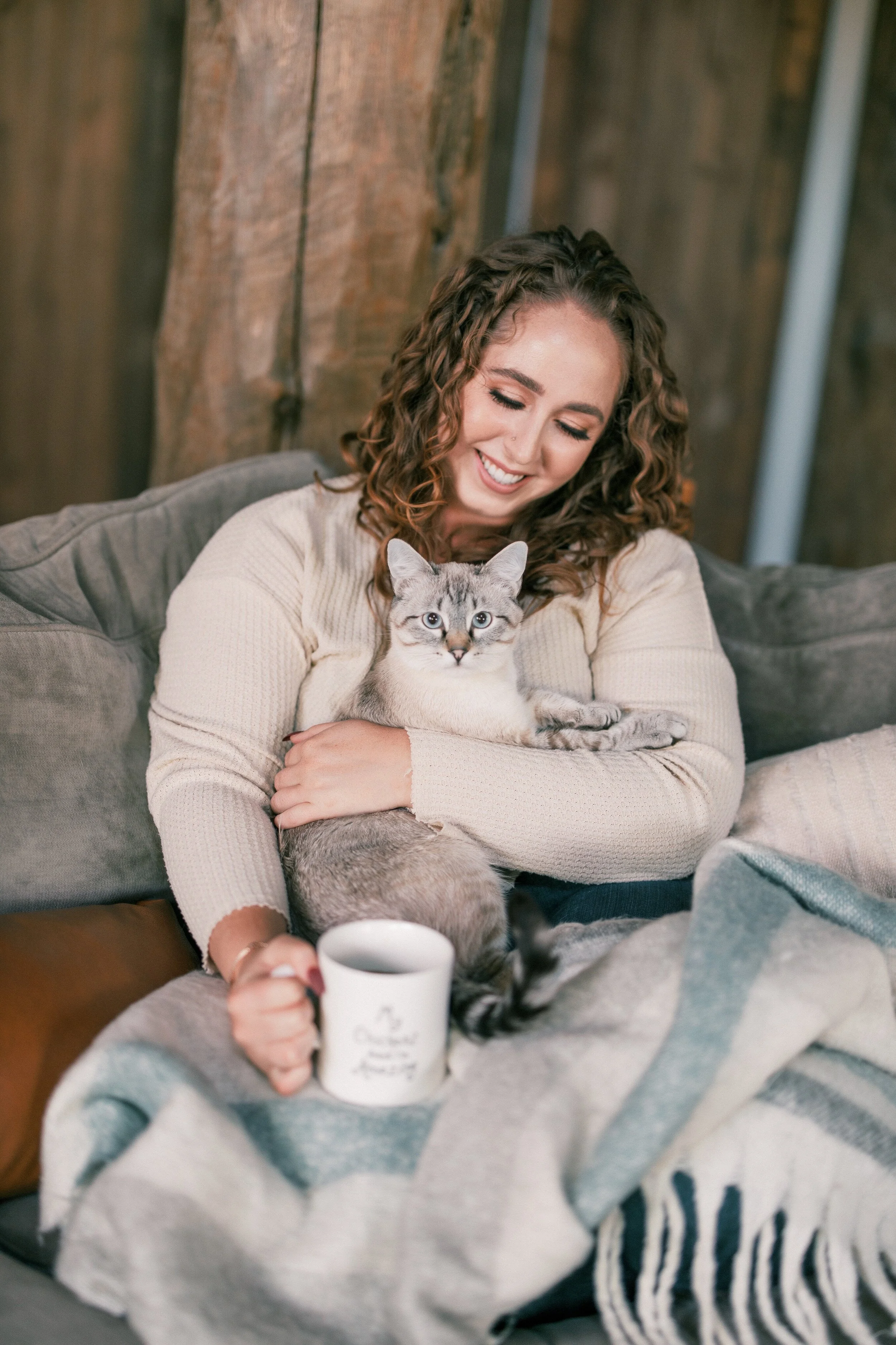 A woman sitting on a sofa holding a gray tabby cat, smiling, with a white mug in her hand, in a cozy living room with wooden walls and a gray blanket with black stripes.