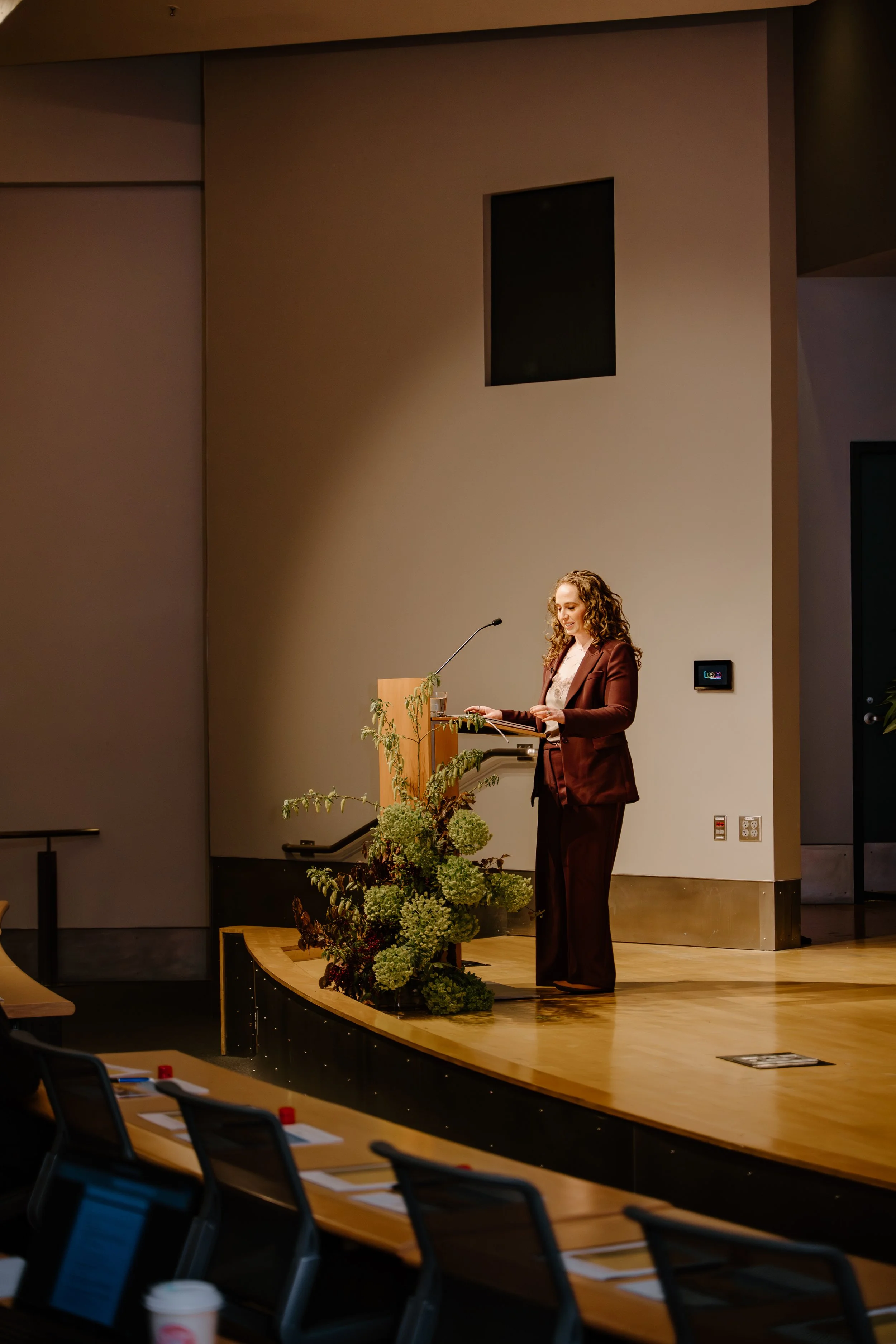 A woman in a brown suit stands at a podium with a microphone in a conference room, smiling. There are potted plants on the stage and audience chairs in the foreground.