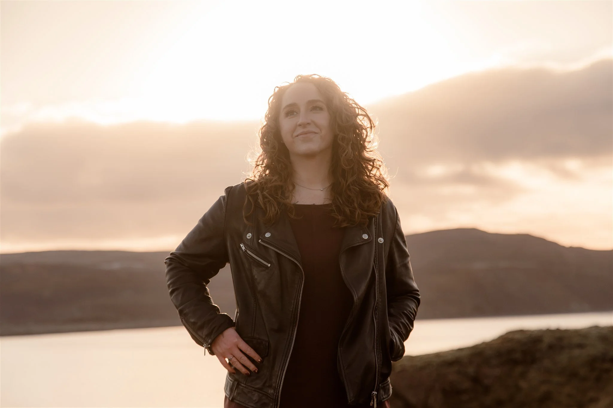 A woman with curly hair wearing a black leather jacket standing outdoors during sunset with hills and a partly cloudy sky in the background.