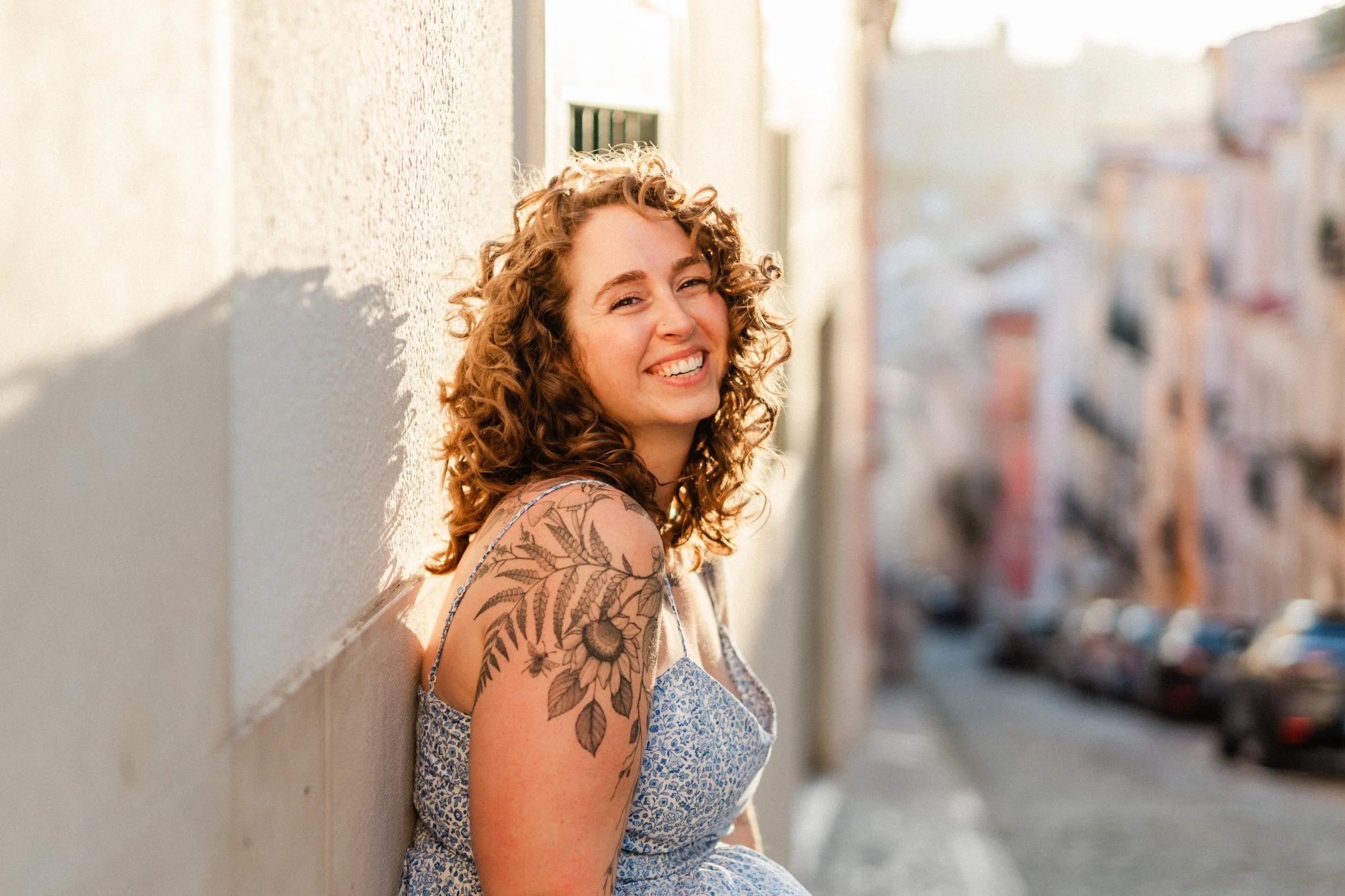 A young woman with curly hair and a floral tattoo on her shoulder is smiling while leaning against a wall in an outdoor urban setting with blurred buildings and street scene in the background.
