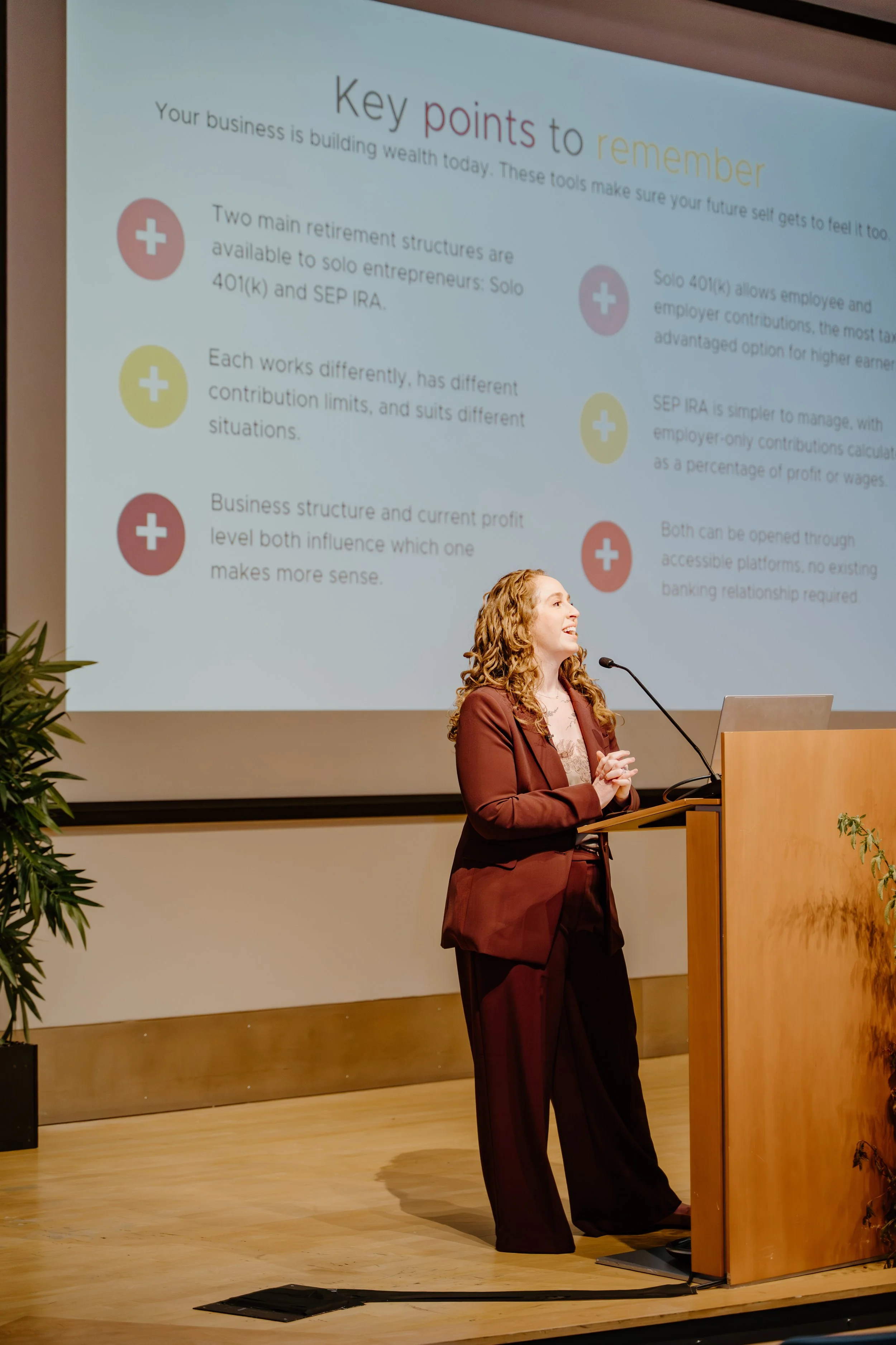 A woman in a brown suit smiling while presenting at a conference. Behind her, a large screen displays key points about retirement plans.