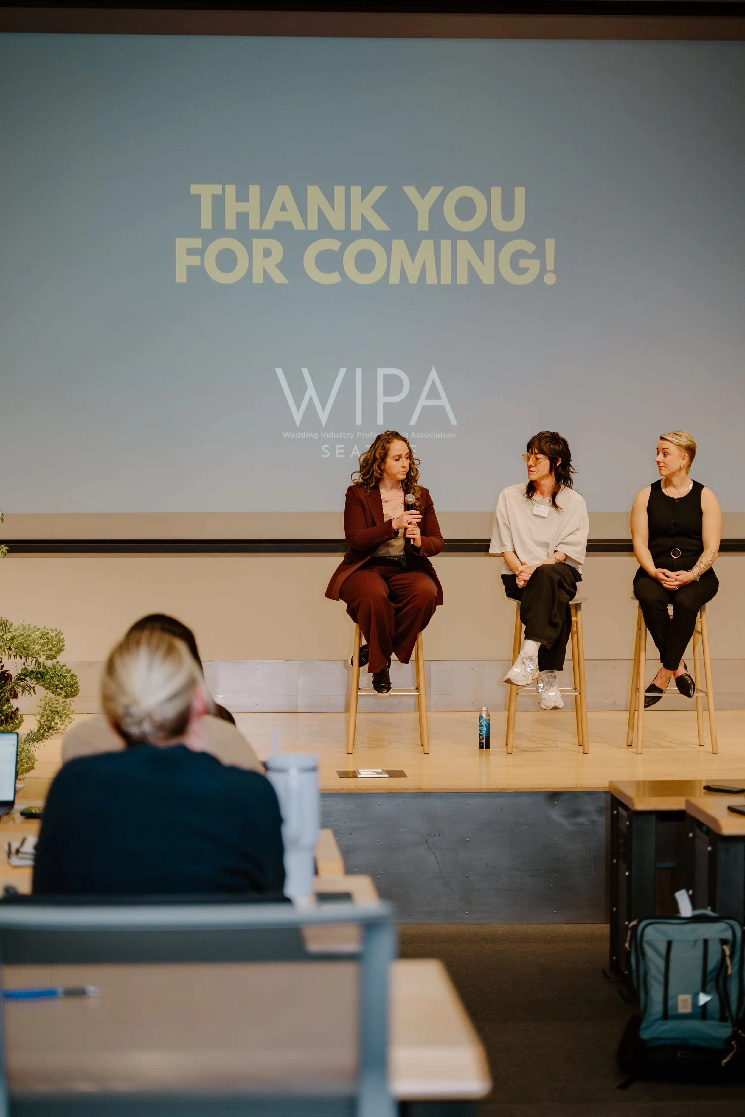 Panel discussion at a conference, with three women sitting on stage in front of a large screen that displays a thank you message for coming from WIPA (Wedding Industry Professionals Association). One woman is holding a microphone, and audience members are visible in the foreground.