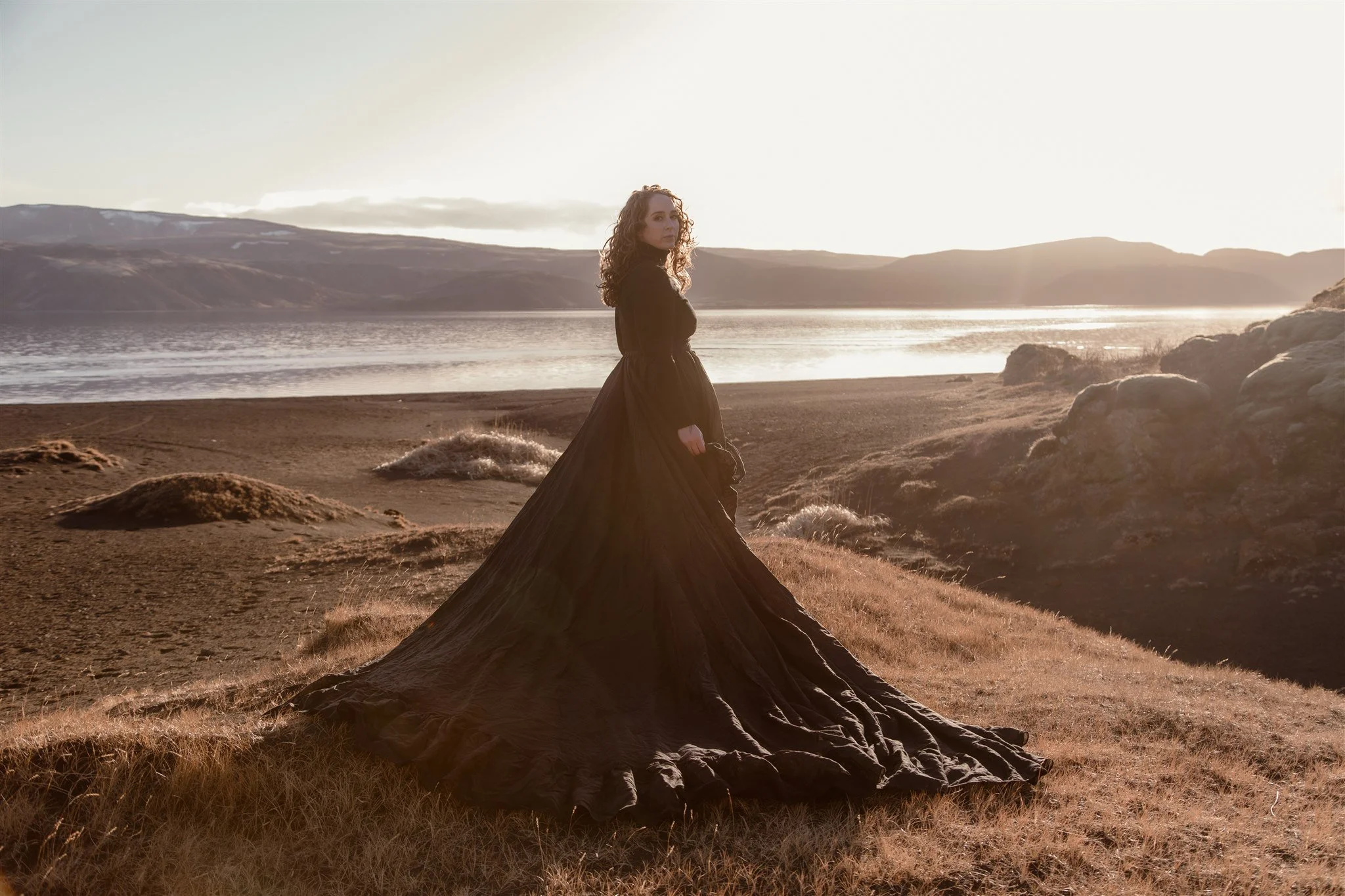 A woman with curly hair wearing a black dress standing on rocky terrain by a lake during sunset.