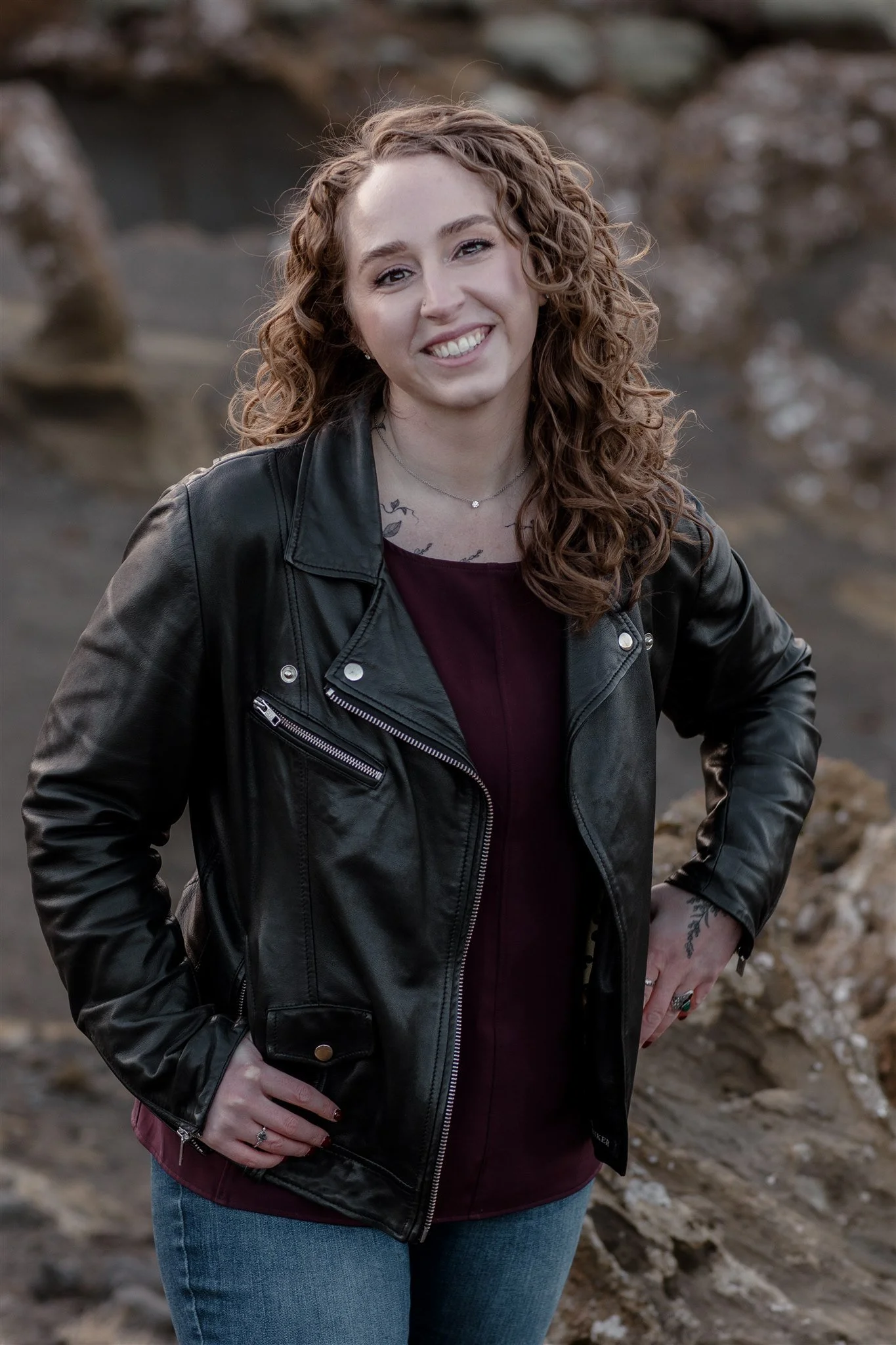 A woman with curly red hair smiling outdoors, wearing a black leather jacket, a maroon top, and blue jeans, standing against a rocky background.