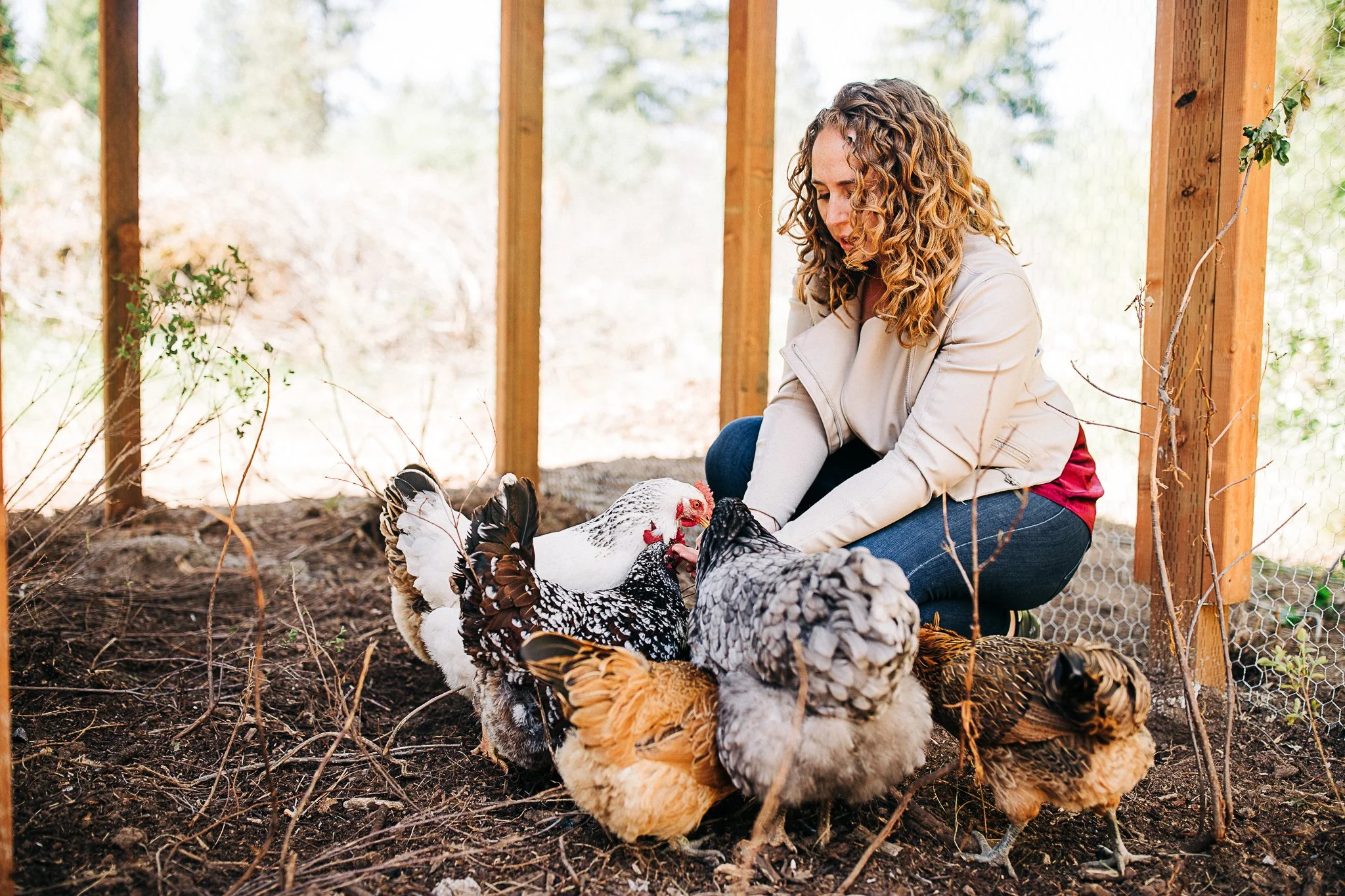 A woman with curly hair wearing a beige jacket and blue jeans is kneeling on the ground inside a fenced chicken coop, feeding and tending to a group of turkeys.
