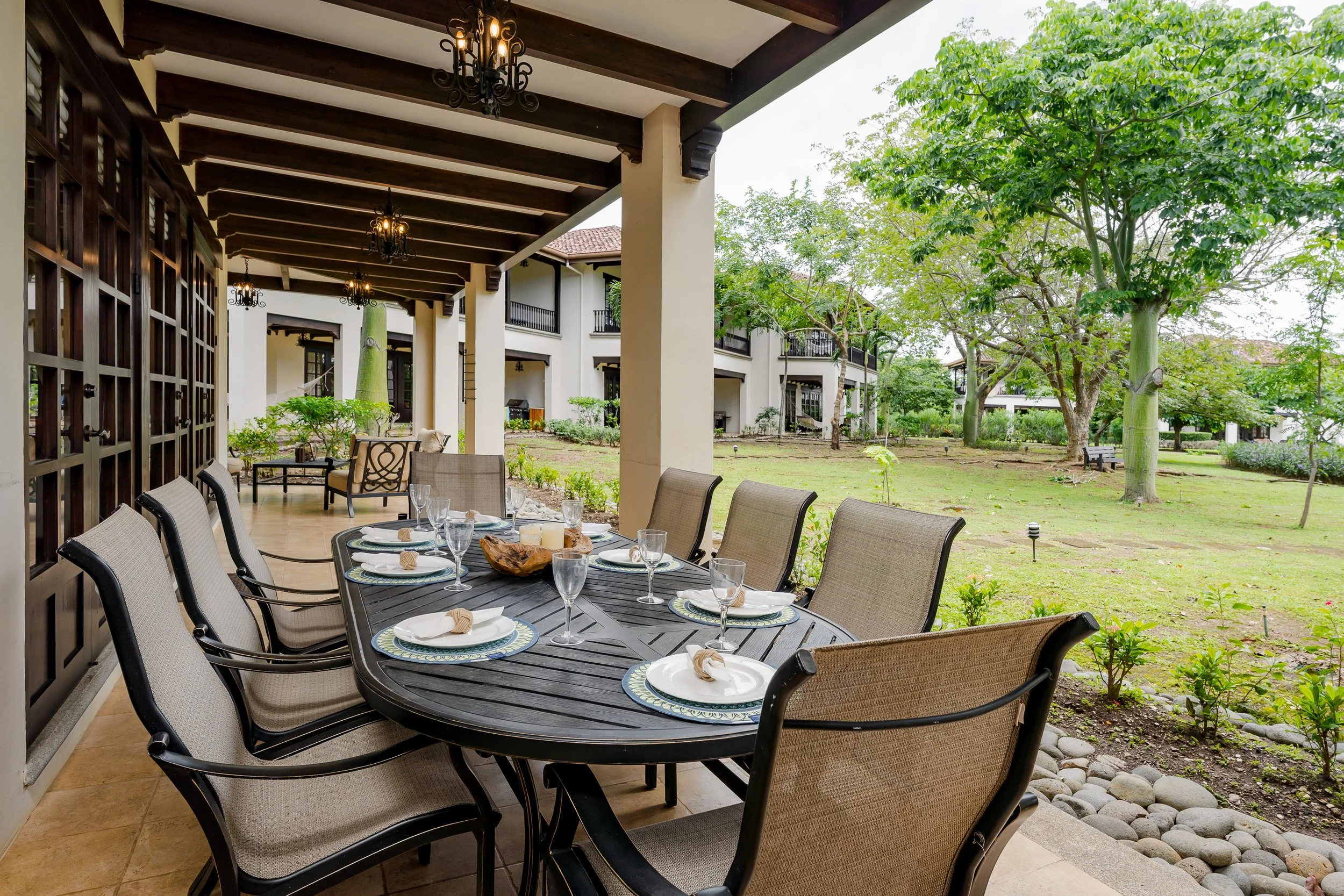 Outdoor dining area with a long table set for a meal, surrounded by beige chairs, with a view of a grassy yard, trees, and multi-story residential buildings in the background.