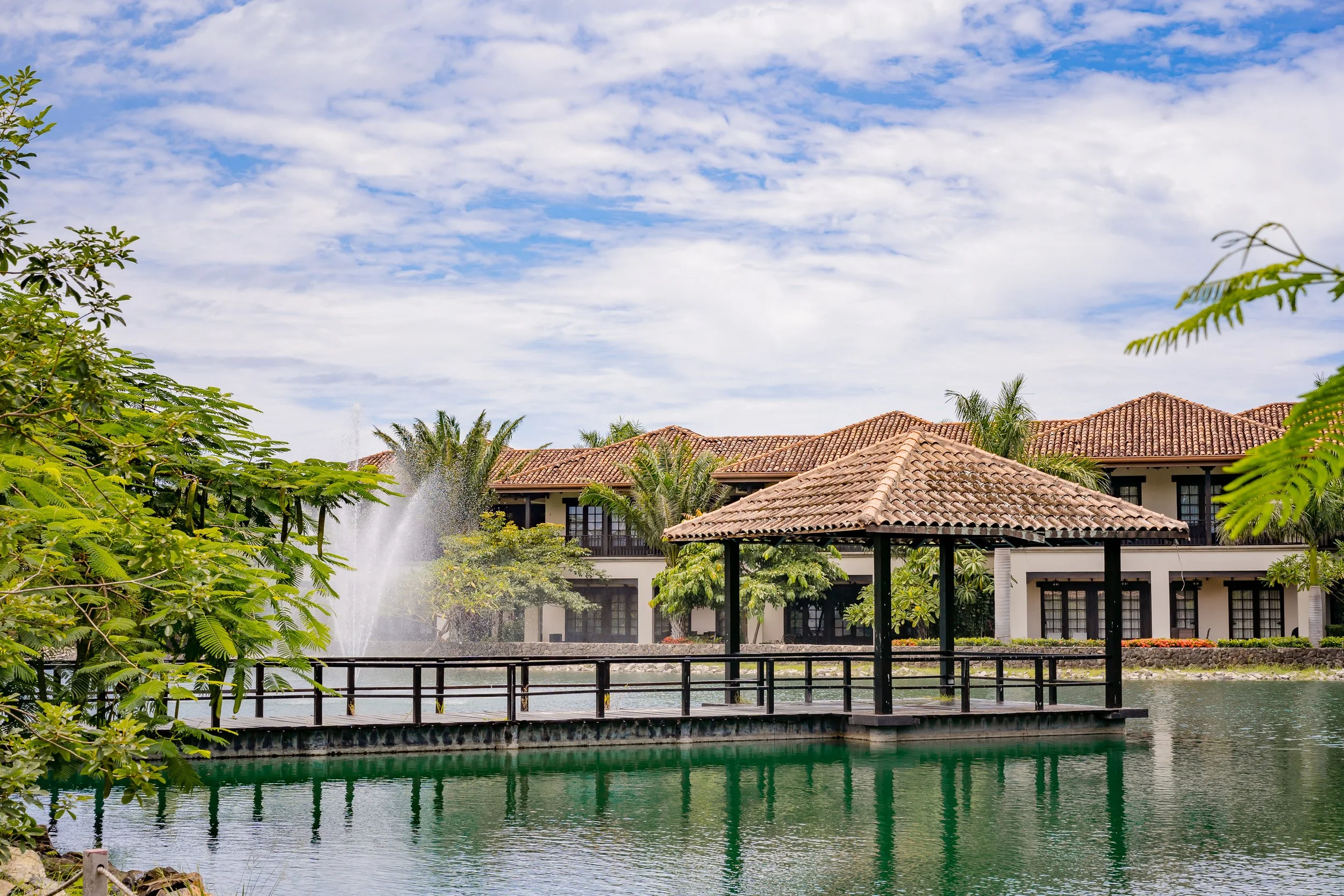 A pond with a wooden walkway and a fountain surrounded by lush green trees and a building with a red tiled roof under a partly cloudy blue sky.
