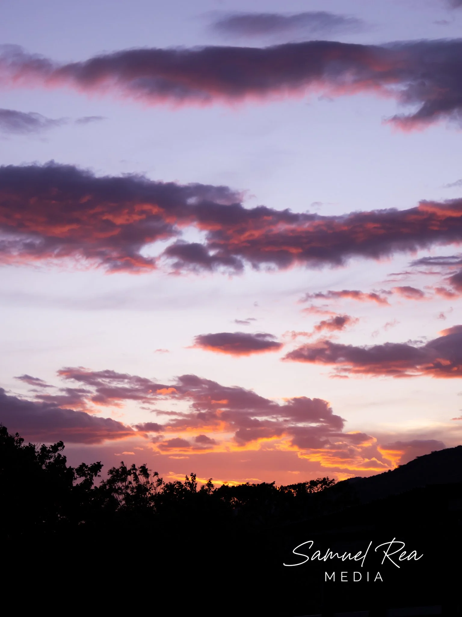 A sunset sky with purple, pink, and orange clouds over a silhouette of trees and hills. The image has a signature text at the bottom right that reads 'Samuel Rea Media'.