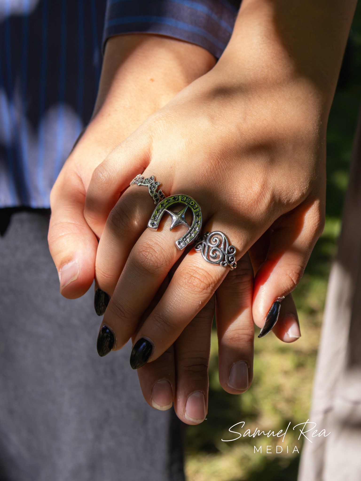 A close-up of two hands intertwined, wearing rings, with a person in a striped shirt in the background. The image is captured outdoors with natural light.