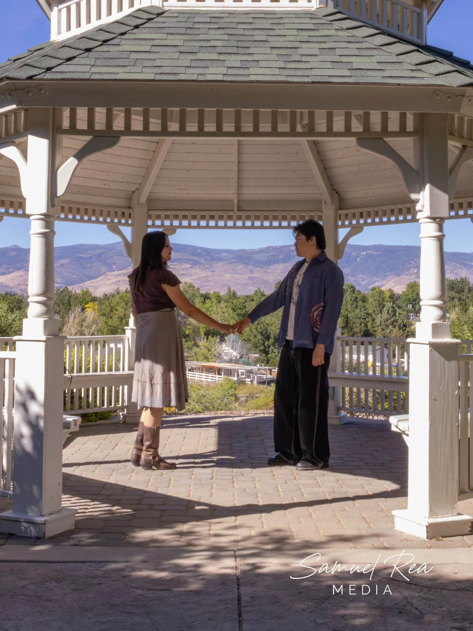 A man and a woman holding hands under a white gazebo with brown shingle roof, overlooking a scenic mountain landscape in the background, during daylight.