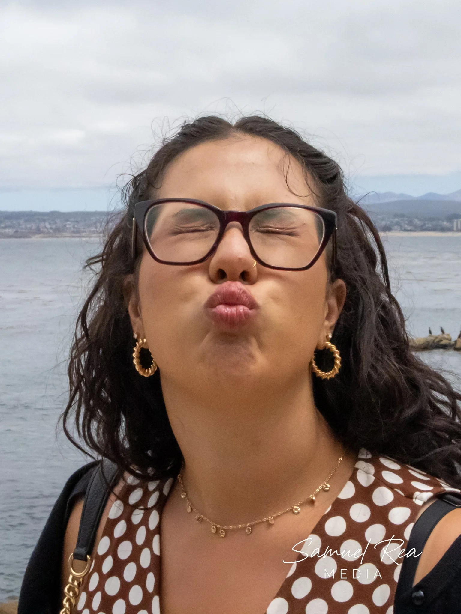 A woman with dark curly hair, wearing glasses, gold earrings, and a gold necklace, making a kissing face with closed eyes. She is dressed in a brown and white polka dot top. Behind her, there is a body of water, mountains, and a cloudy sky.