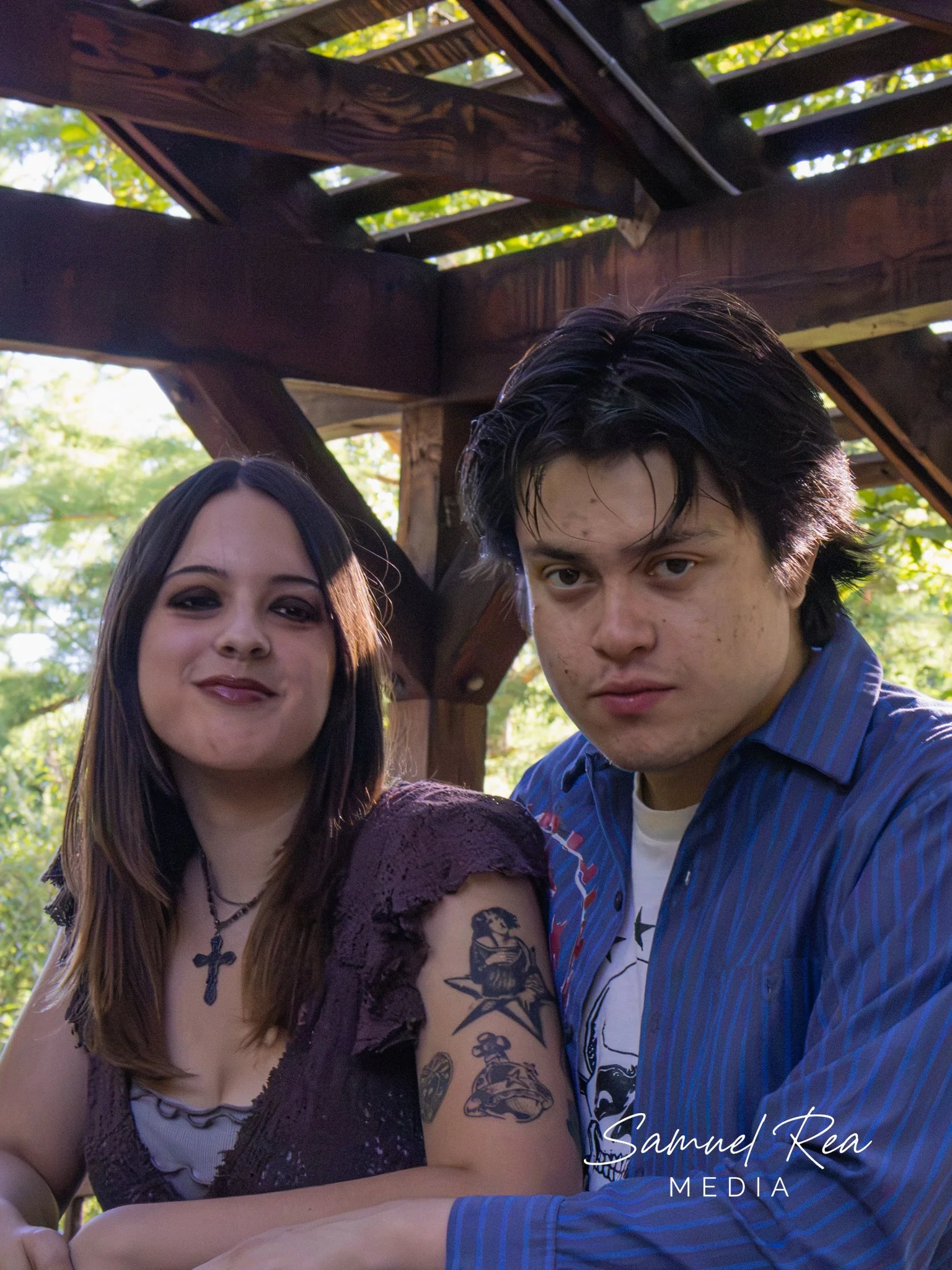 Two people are standing close together outdoors with a wooden structure and trees in the background. The person on the left is a woman with long brown hair, wearing a purple lace top and a cross necklace. The person on the right is a man with black, 
