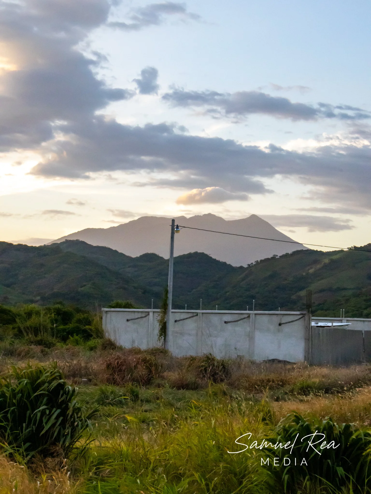 Scenic view of green hills at sunset with a mountain in the background, a cloudy sky, and a utility pole in the foreground. Text overlay reads 'Samuel Rea Media'.