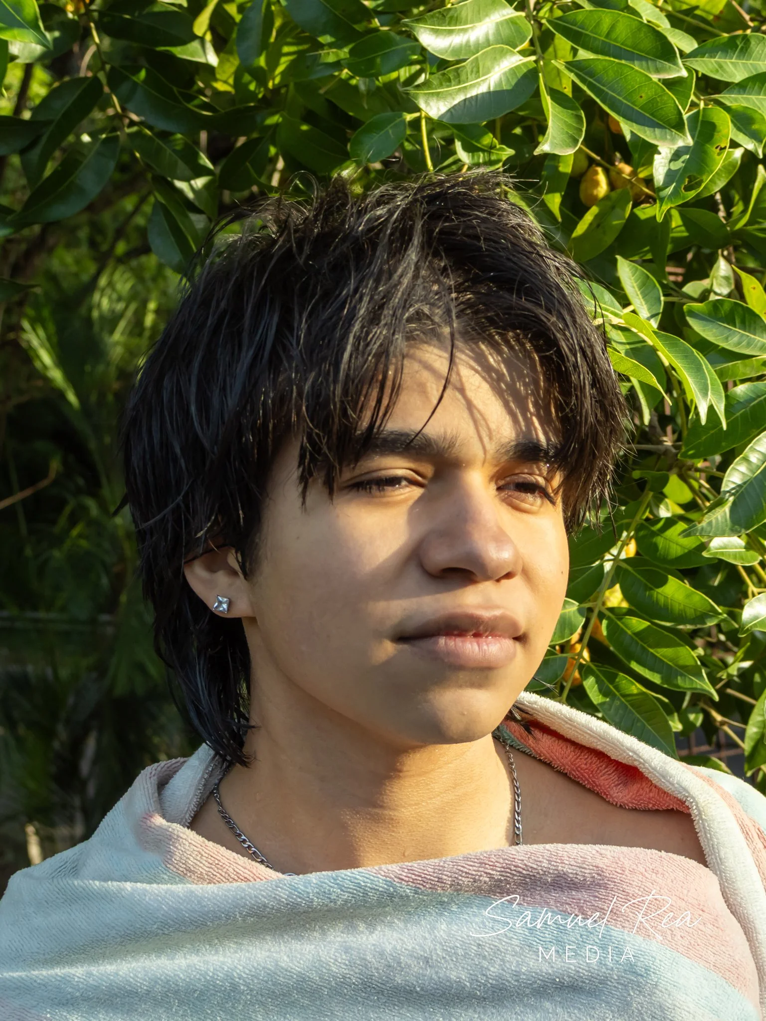 Close-up of a young person's face with wet dark hair, wearing earrings and a chain necklace, outdoors with green leafy plants in the background, sunlight illuminating their face.