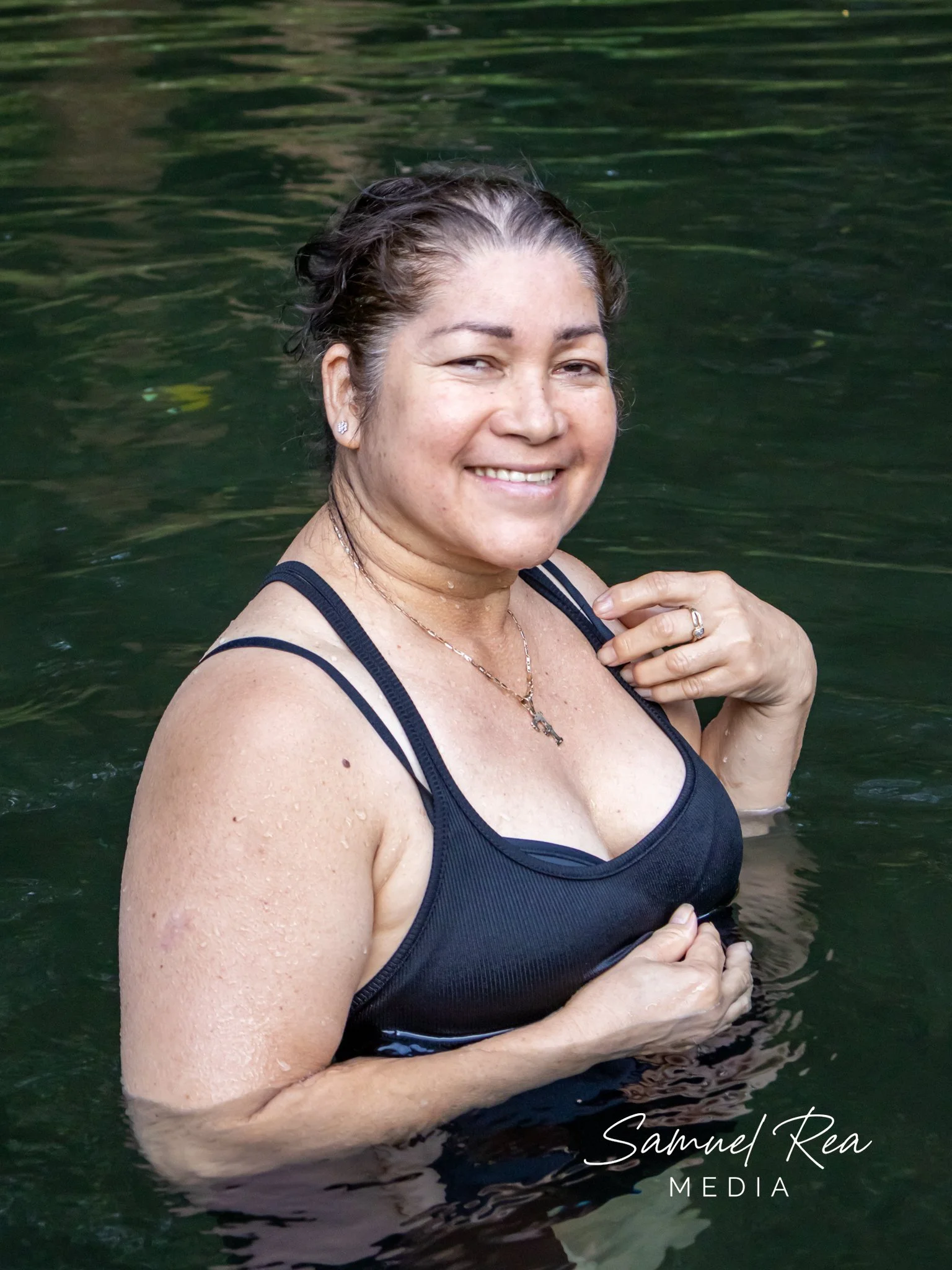 A woman with wet hair, wearing a black top and jewelry, smiling and standing in a body of water, with her hand resting on her shoulder, partially submerged. The water appears to be greenish, and the background is blurred water.
