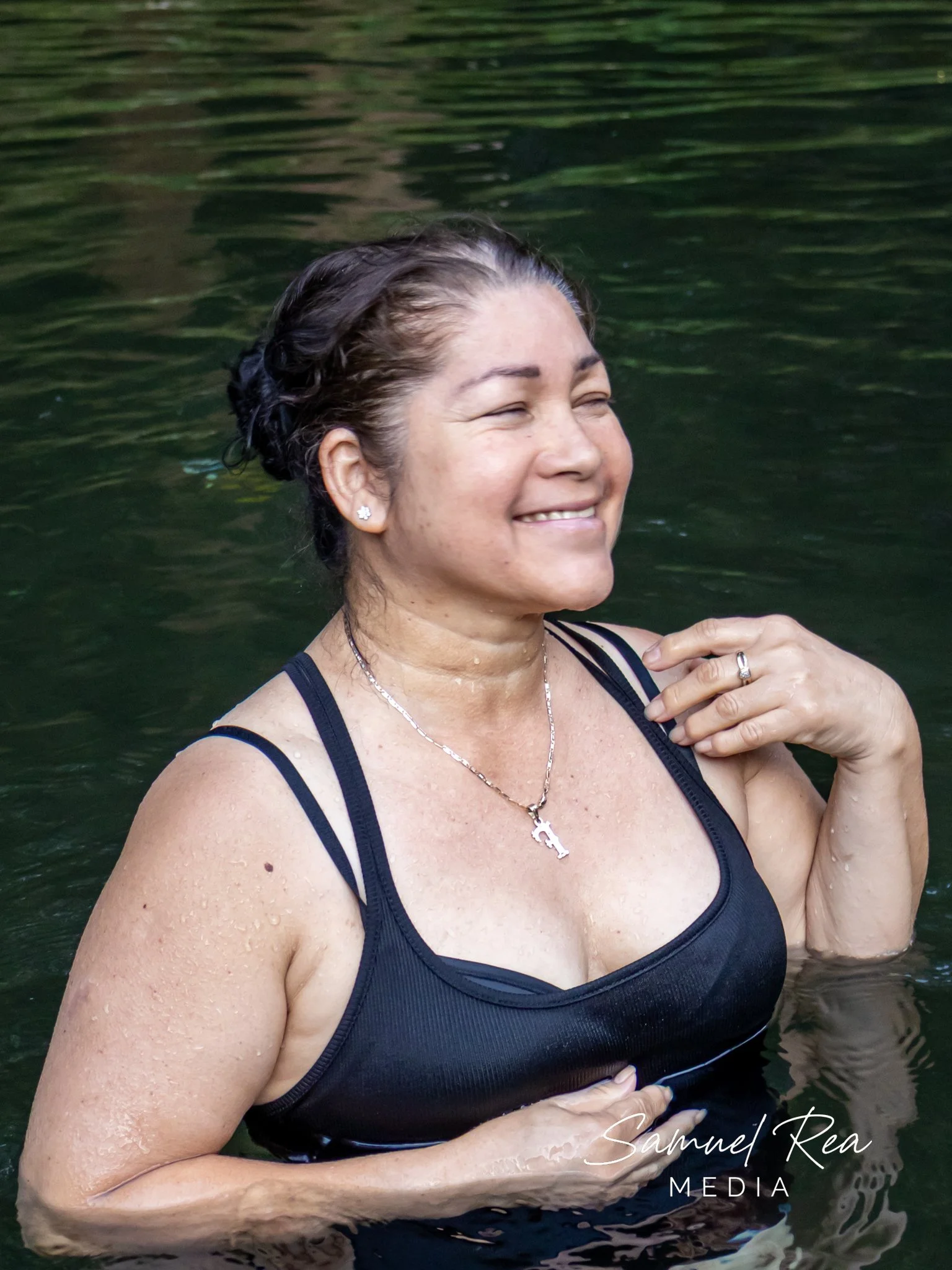 A woman with wet hair smiling in a body of water, wearing a black tank top and a cross necklace, with her hand resting on her shoulder, during daytime.