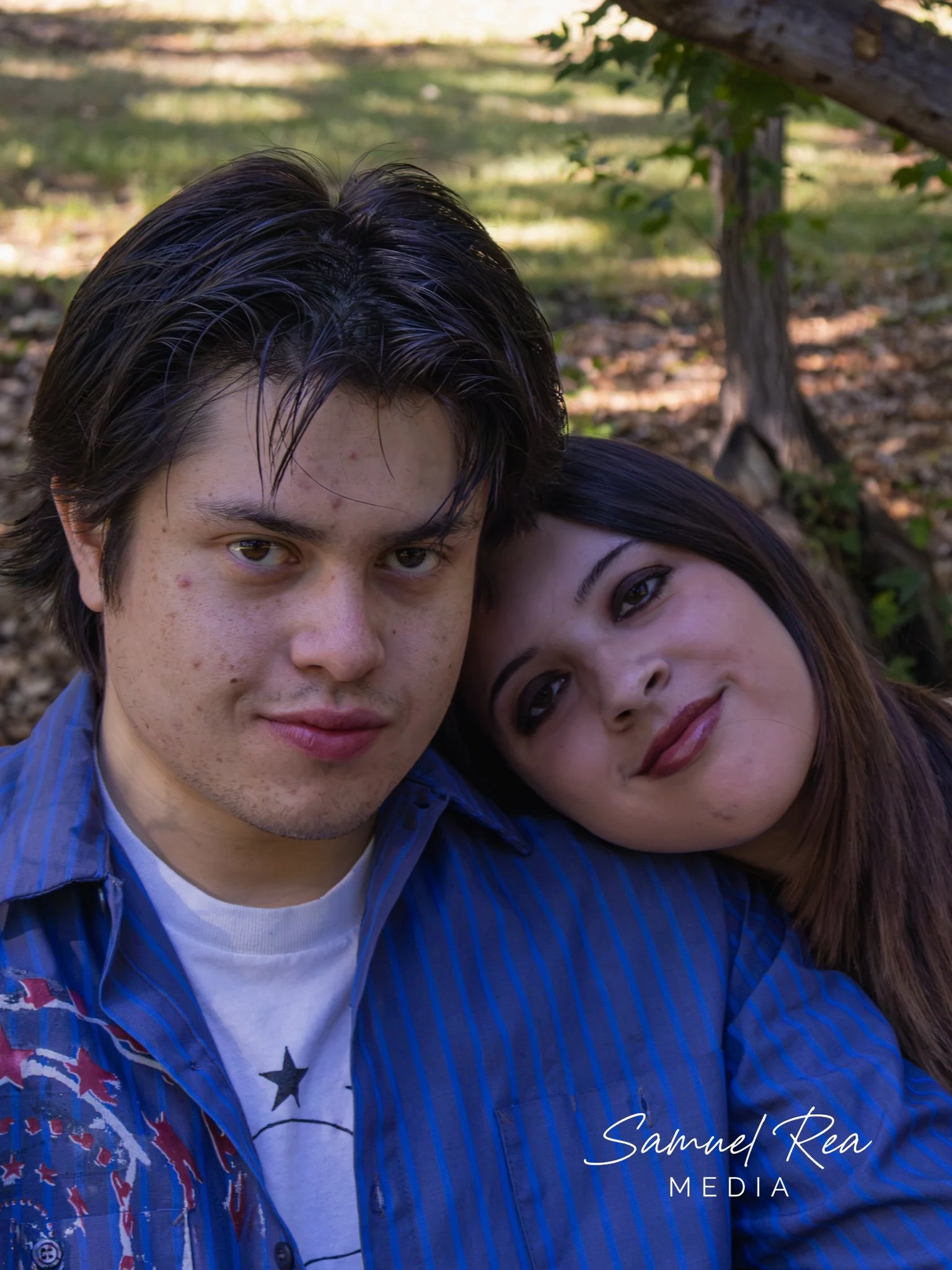 A young man and woman close together outdoors, with trees and leaves in the background, smiling at the camera.