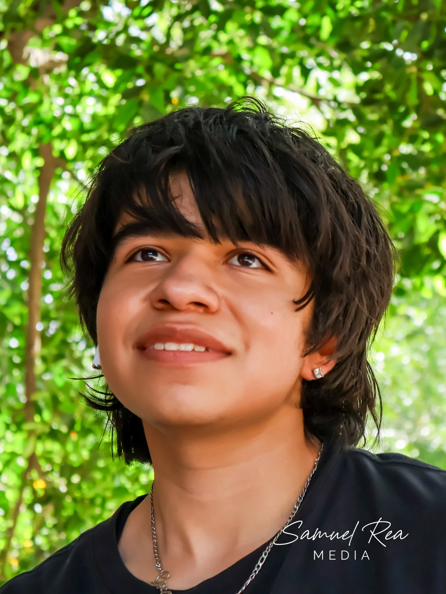 Close-up of a smiling person with short dark hair looking upward, wearing earrings and a necklace, with a green leafy background.