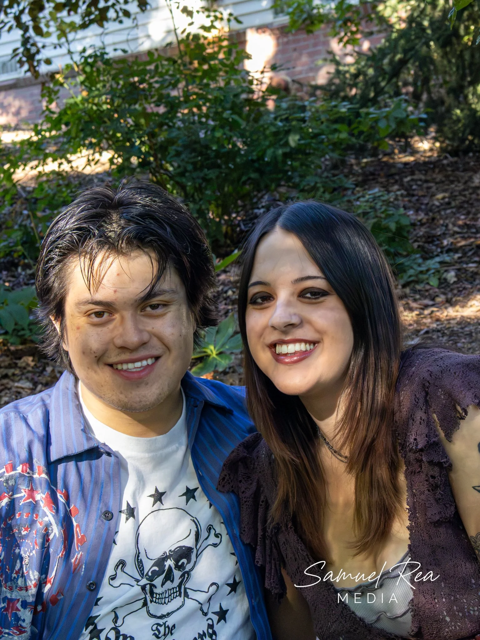 A smiling young man and woman taking a selfie outdoors with greenery and a brick building in the background.
