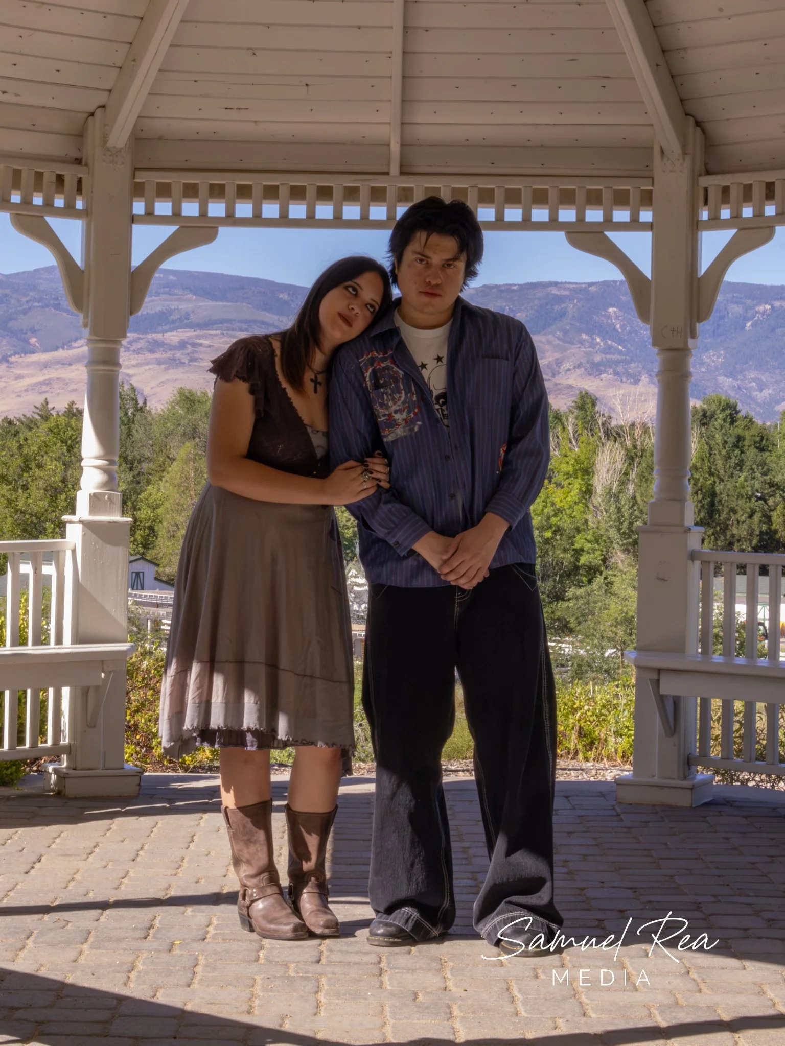 A man and a woman standing close together under a gazebo, with mountains and trees in the background.