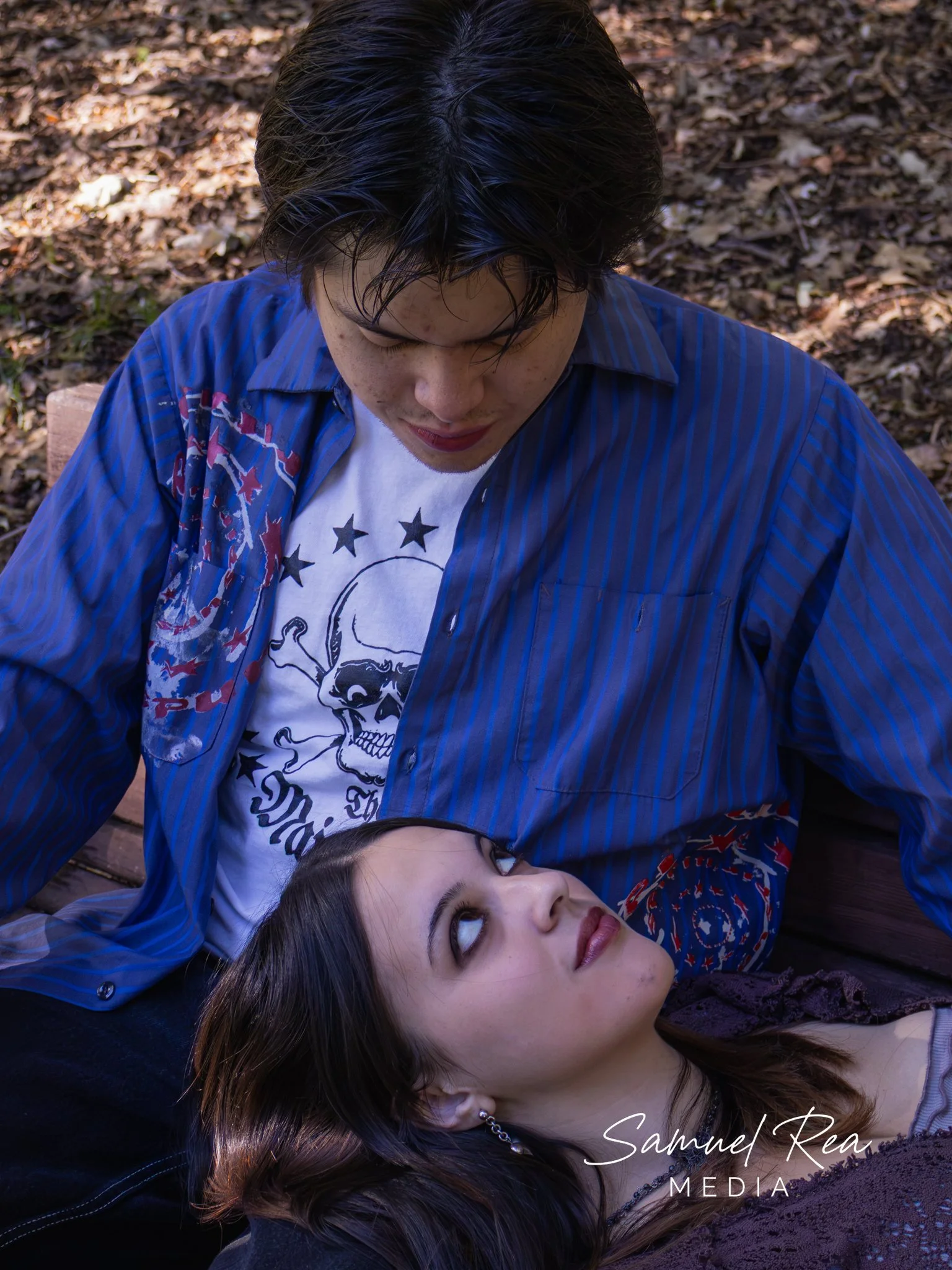 A young woman with long dark hair lying on the ground and a young man with dark hair sitting above her in a close-up outdoor setting with fallen leaves. They are looking at each other, and the woman's head is tilted back. The image has a signature th