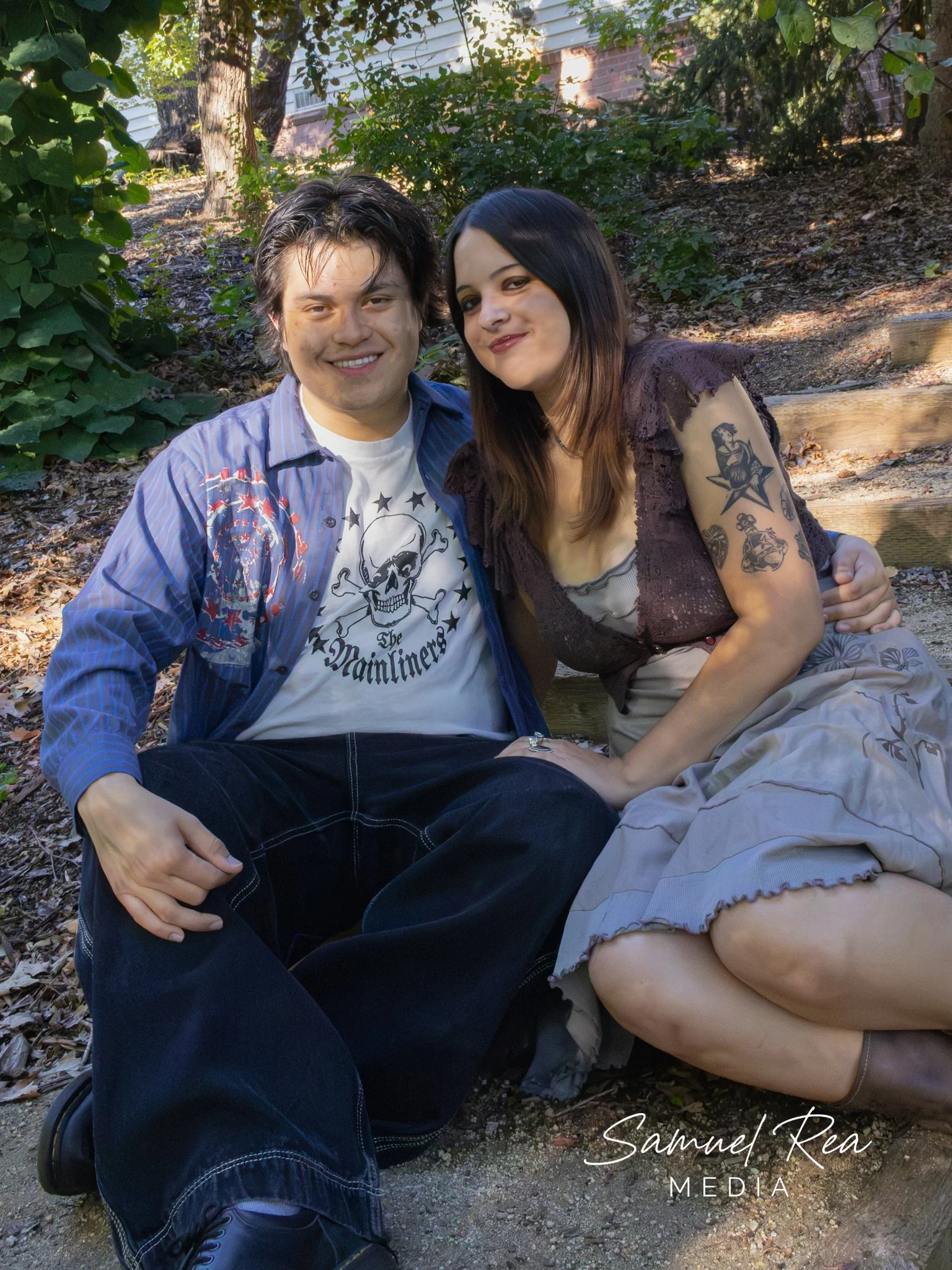 A young man and woman sitting outdoors on the ground, surrounded by trees and plants, smiling at the camera. The man has dark, slightly wet hair, a shirt with a skull logo, and dark pants. The woman has long dark hair, tattoos on her right arm, and i