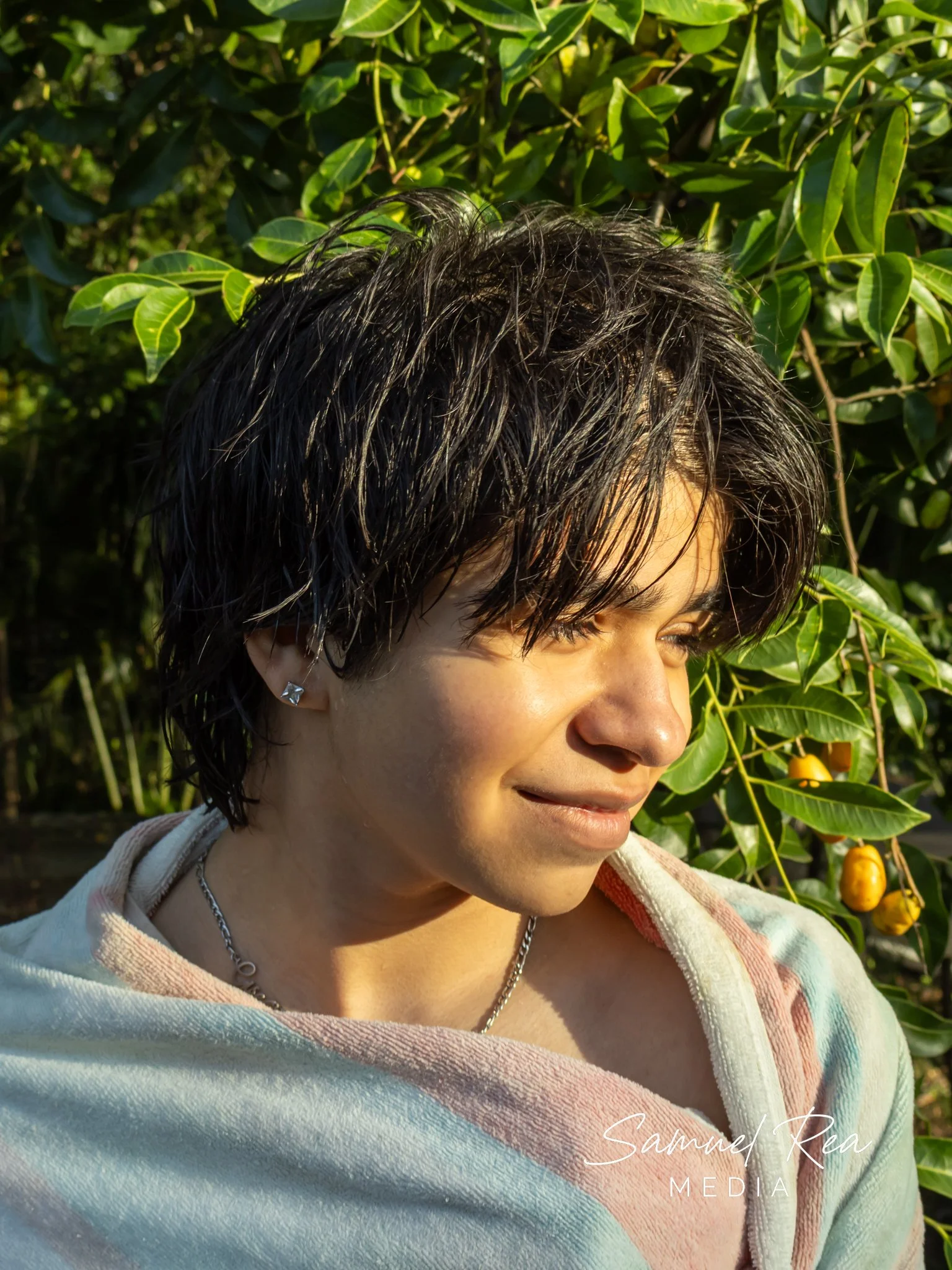 A woman with short black hair and earrings standing outdoors in front of green leafy plants with yellow fruit, wearing a multicolored hoodie and silver necklace, smiling slightly.
