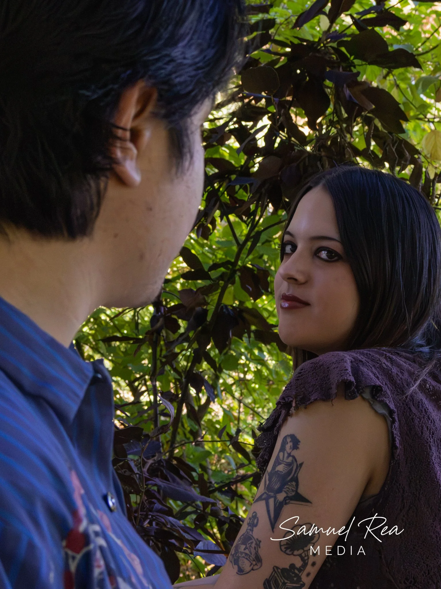 Close-up of a man and a woman looking at each other among green leaves. The woman has long black hair, tattoos on her arm, and is wearing a purple lace top. The man has dark hair and is wearing a blue striped shirt.