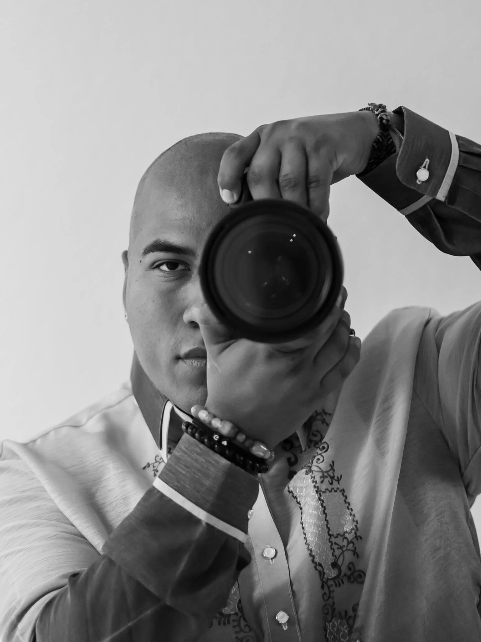 A person taking a selfie in a mirror, holding a camera lens in front of their face. The person is wearing a traditional embroidered shirt with decorative band on the sleeve and a necklace.