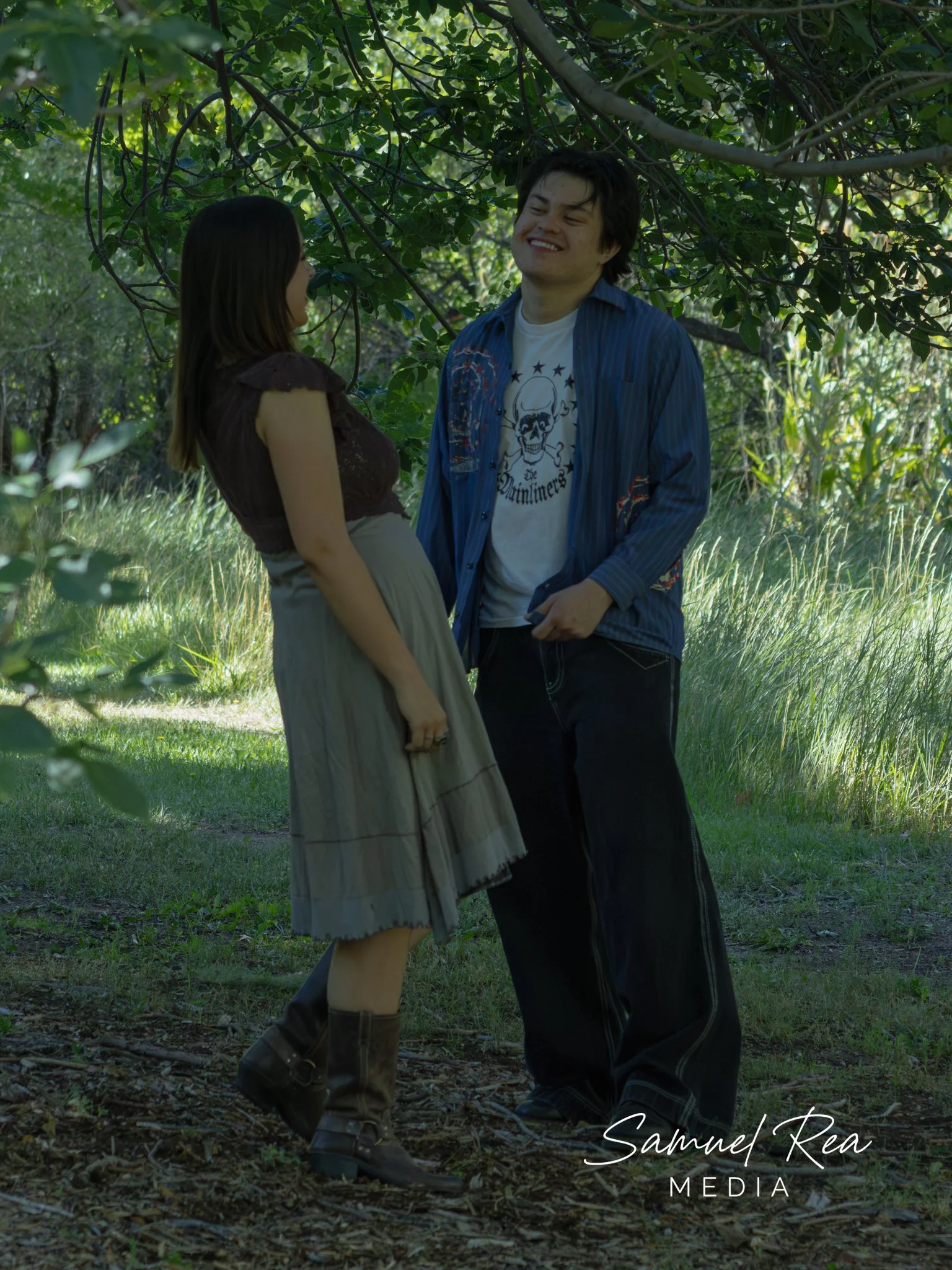 A young man and young woman standing close together in a lush green outdoor setting, smiling at each other. The woman is dressed in a brown top and beige skirt, while the man is wearing a blue jacket over a graphic T-shirt and black pants. The backgr