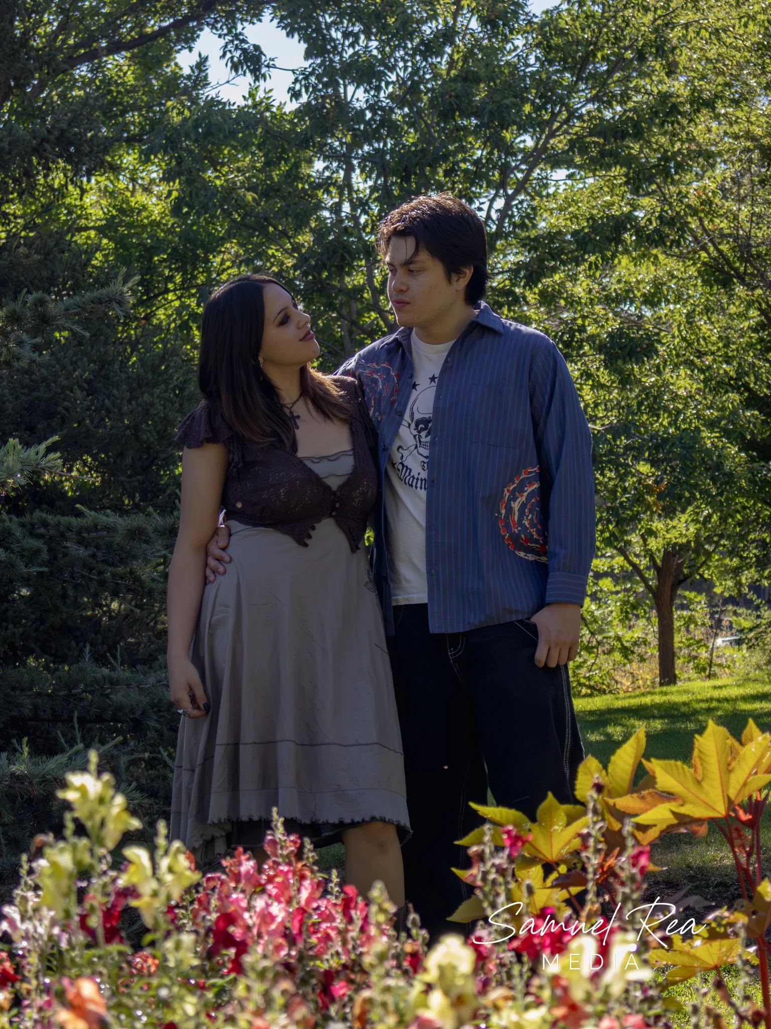 A young couple with light skin stands outdoors among lush green trees and colorful flowers. The woman has dark hair and wears a brown dress, while the man has dark hair and wears a blue striped shirt over a white graphic T-shirt. They are close, gazi