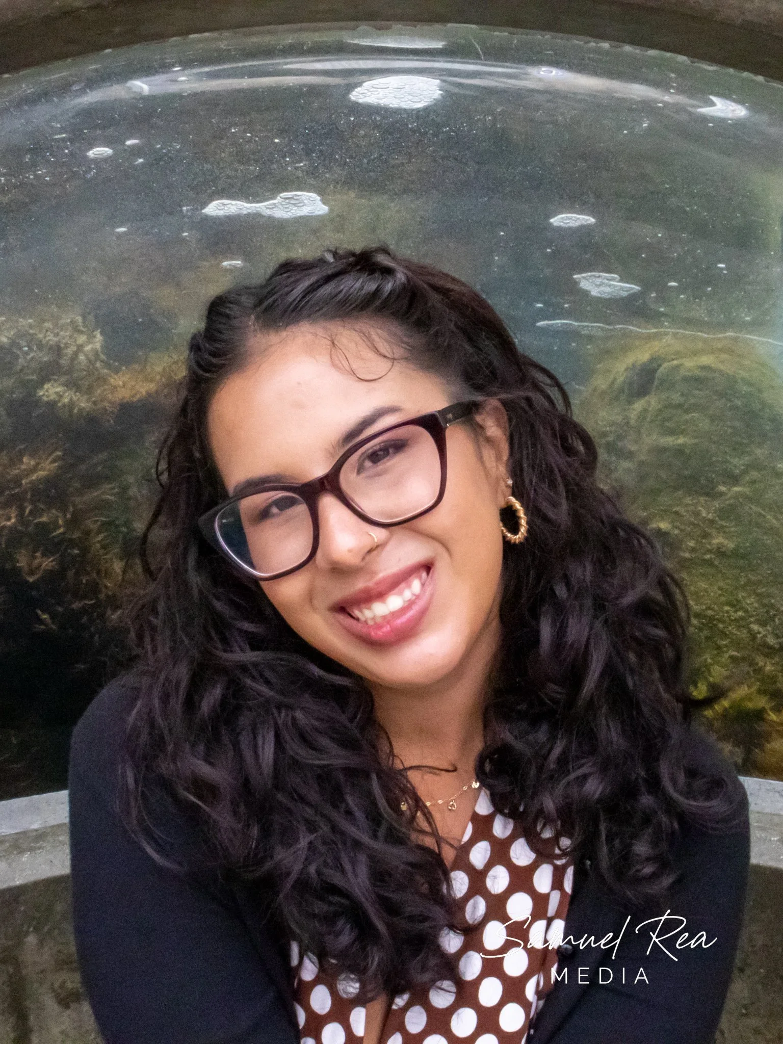 A woman with curly dark hair wearing glasses, gold hoop earrings, a gold nose ring, a necklace, and a black cardigan, smiling in front of a pond or lake with rocks and floating bubbles.
