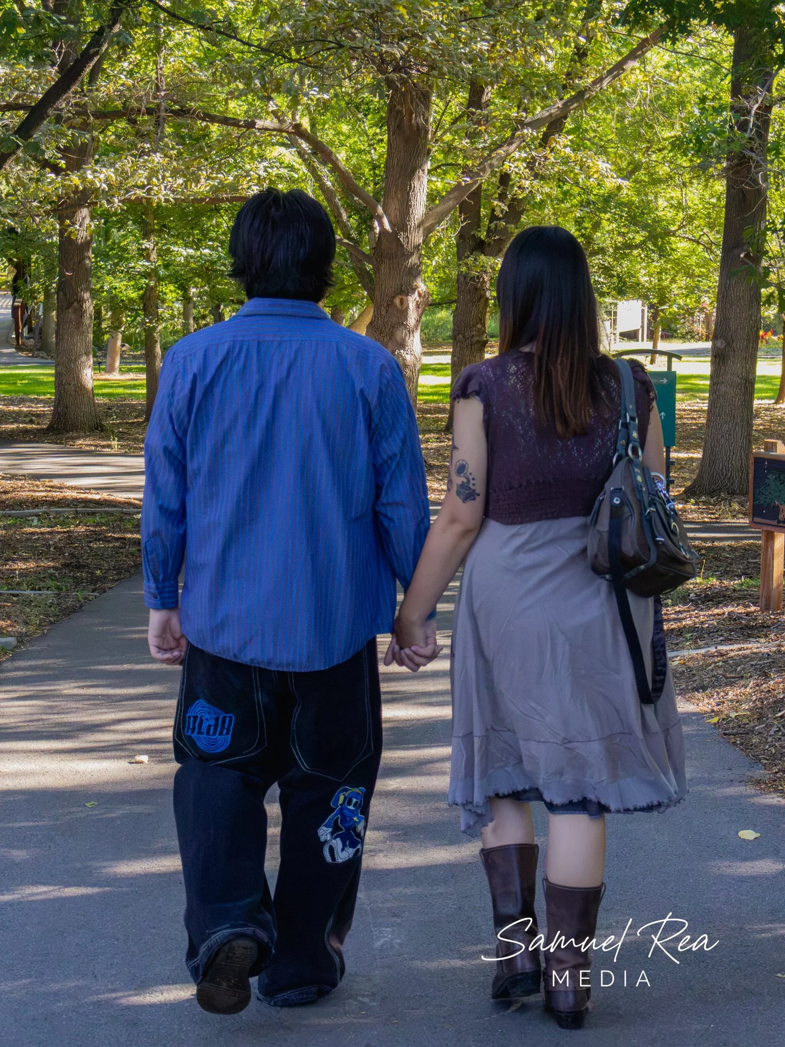 A couple holding hands walking in a park with tall trees and green foliage. The man is wearing a blue shirt and black pants, and the woman is dressed in a purple top with a short skirt and knee-high boots. The sunlight filters through the leaves, cre