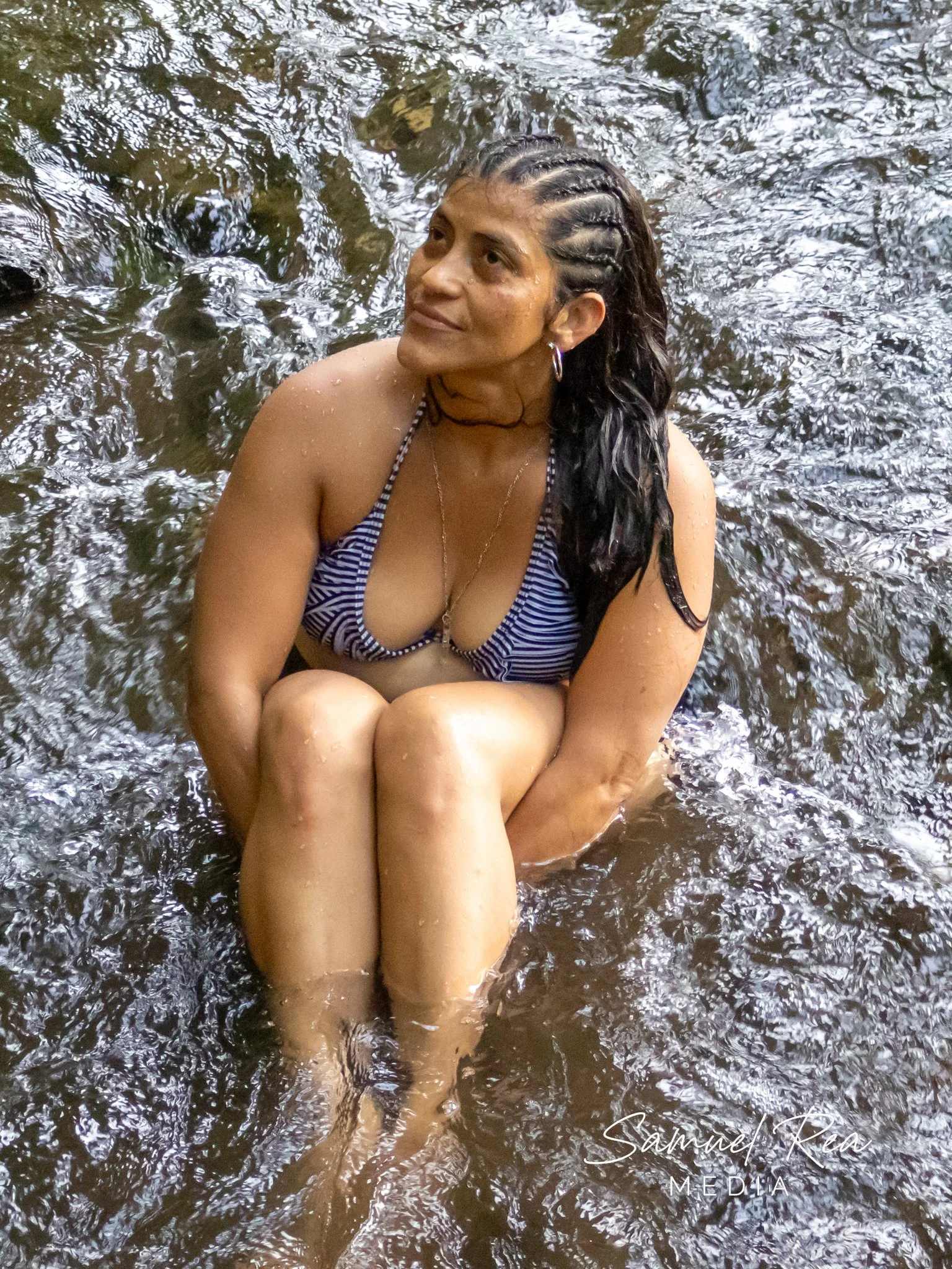 A young woman with braided hair, earrings, and a striped bikini top sitting in a body of water, smiling and looking into the distance.