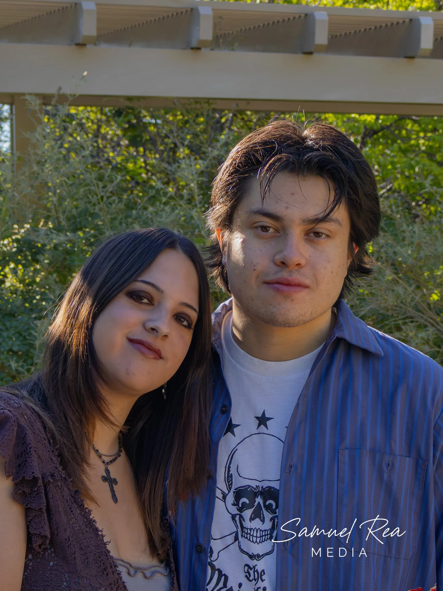 Close-up photo of a woman and a man outdoors with green foliage and a wooden structure in the background. The woman has long dark hair, makeup, and is wearing jewelry, including a cross necklace. The man has short dark hair, some facial blemishes, an