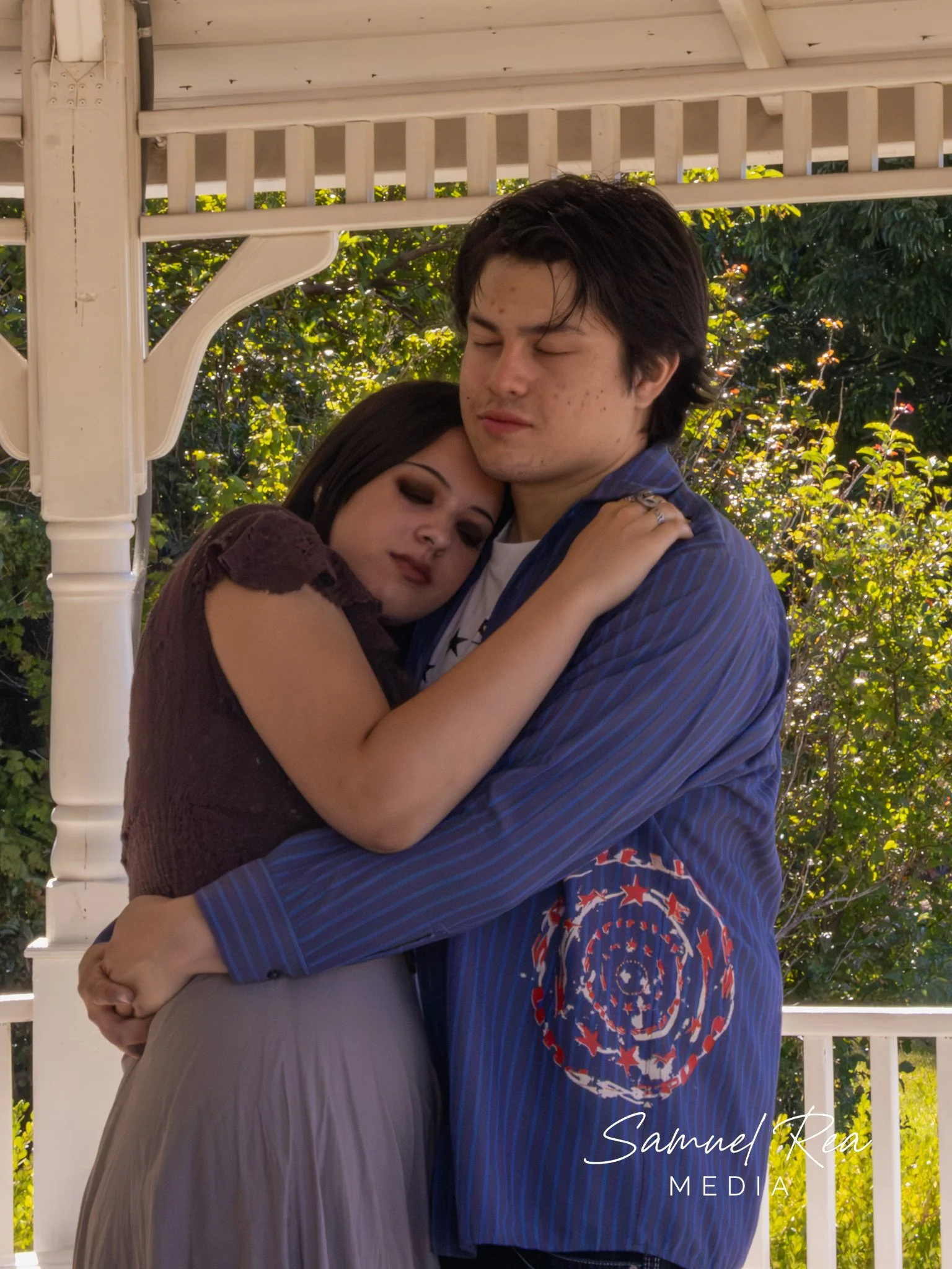 A young couple embracing each other outdoors near a white gazebo, with trees and greenery in the background. The woman rests her head on the man's shoulder, and they both have a calm expression.