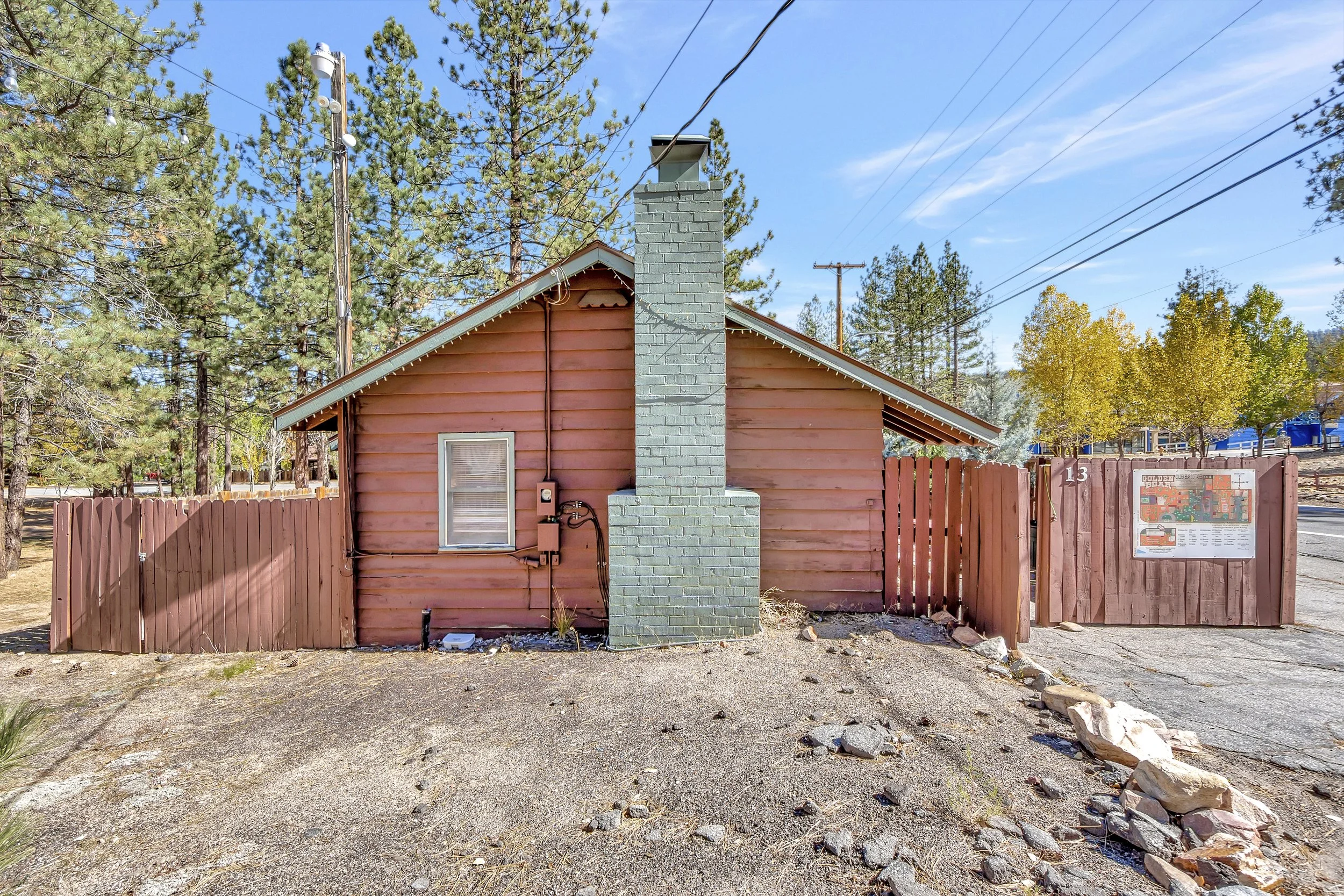 A small pink house with a blue chimney, red wooden fence, surrounded by trees and electrical wires under a blue sky.