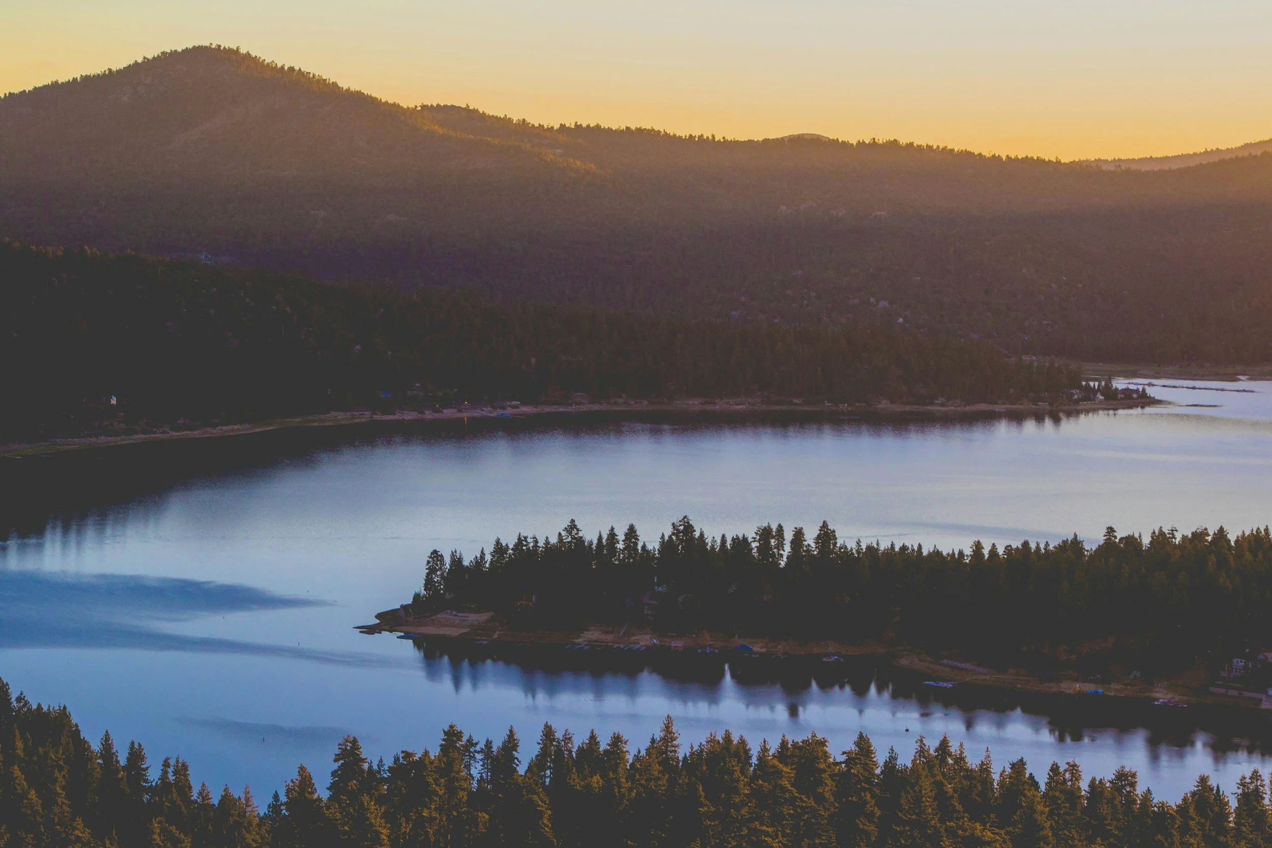 Scenic view of a mountain landscape with a calm body of water in the foreground, surrounded by dense trees, during sunset or sunrise.