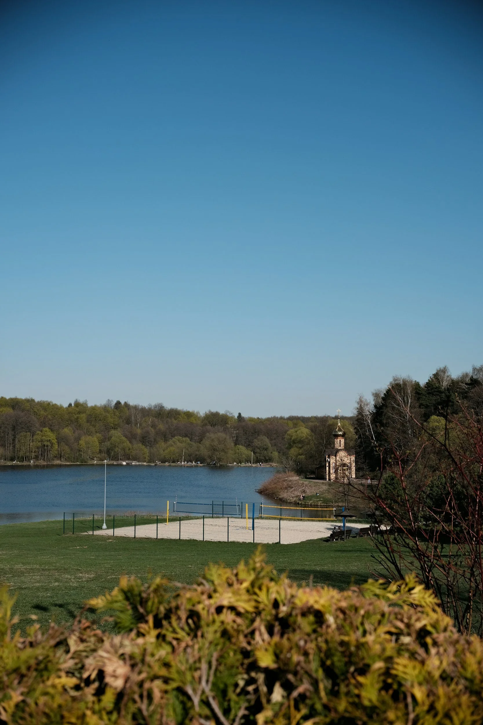 A lakeside park with a grassy area, benches, and a volleyball court, overlooking a calm lake. There is a small church with a golden dome on the right side near trees. The background has a wooded hill and a clear blue sky.