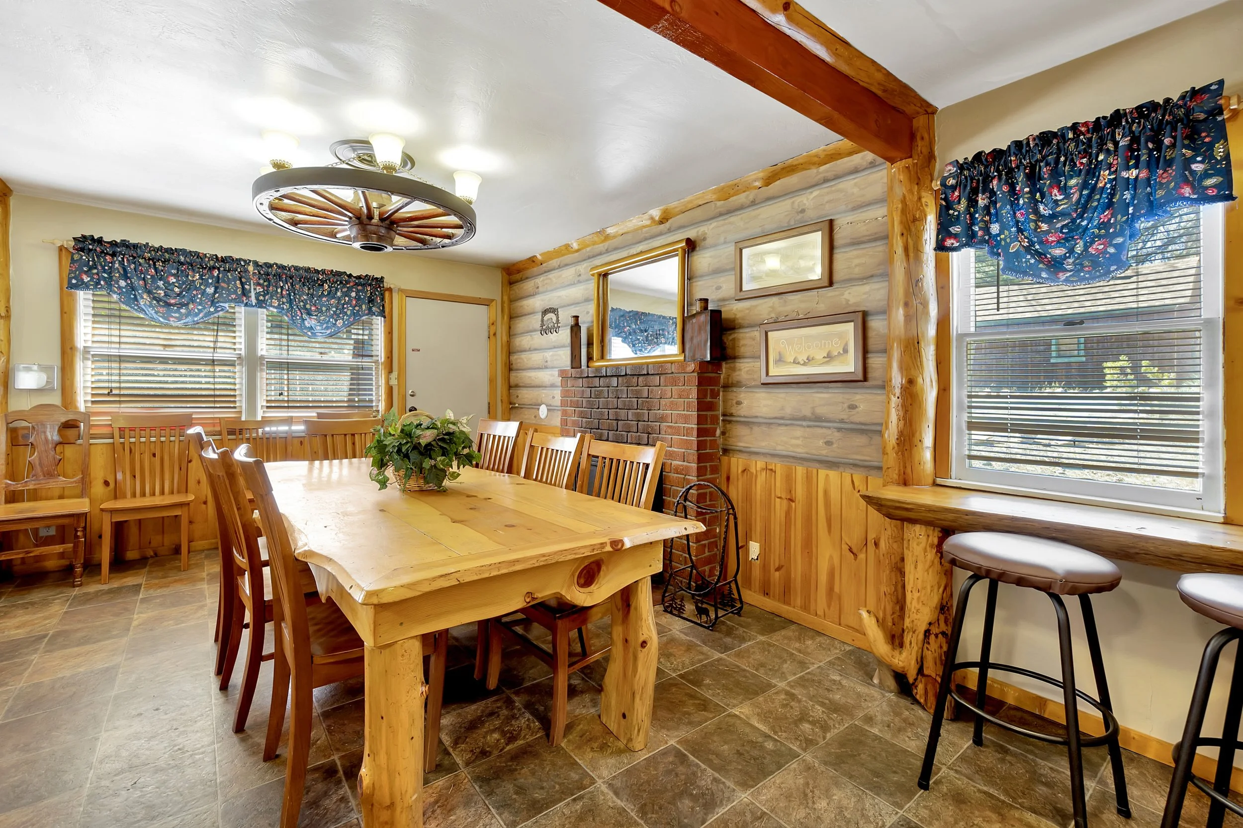 A rustic dining room with wooden log walls, a large rectangular wooden dining table surrounded by matching wooden chairs, a brick fireplace with a mirror above it, and windows with floral valances and white blinds. There are also bar-height stools at