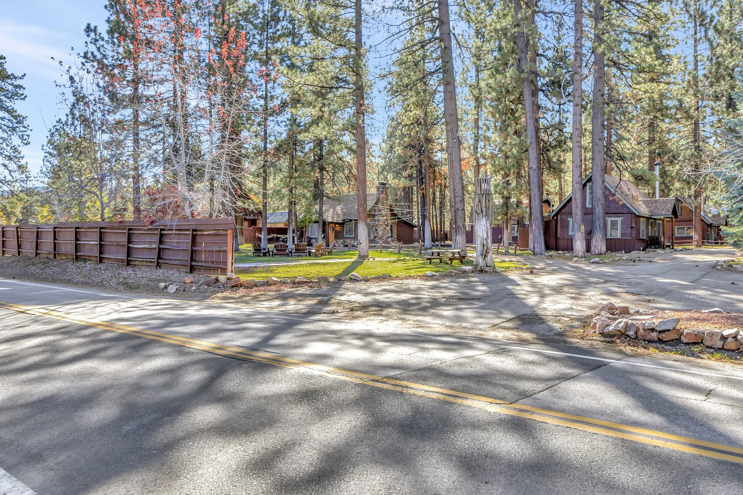 Scenic view of a wooded area with tall pine trees surrounding rustic cabins with wooden exteriors and stone Chimney, outdoor seating including picnic tables and Adirondack chairs, a wooden fence, and a dirt path under a clear blue sky.