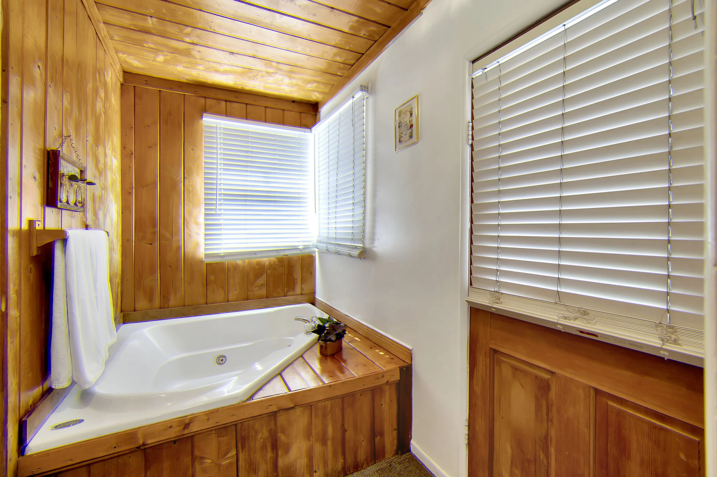 Bathroom with wood-paneled walls and ceiling, white bathtub, two white towels on a wooden towel rack, two windows with white blinds, and a small framed picture on the wall.