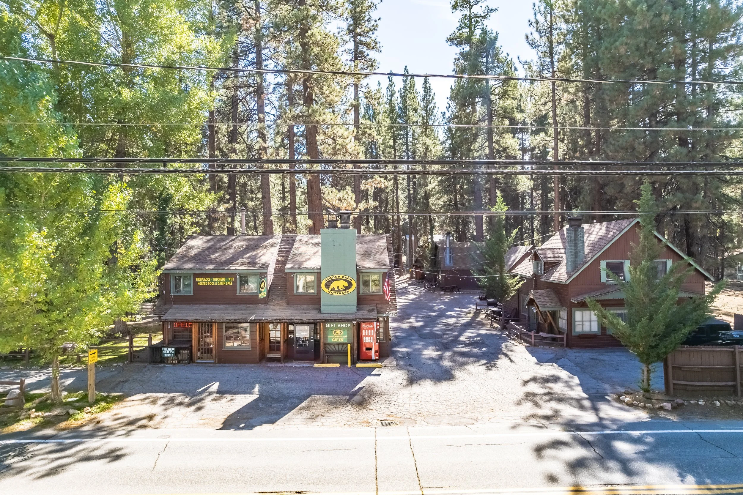A small building with a sign that says "Golden Bear Cottages" and advertisements for fireplaces, kitchens, WiFi, heated pools, and cabin spas. There are trees and houses surrounding the area, with electrical wires overhead and a paved parking lot.